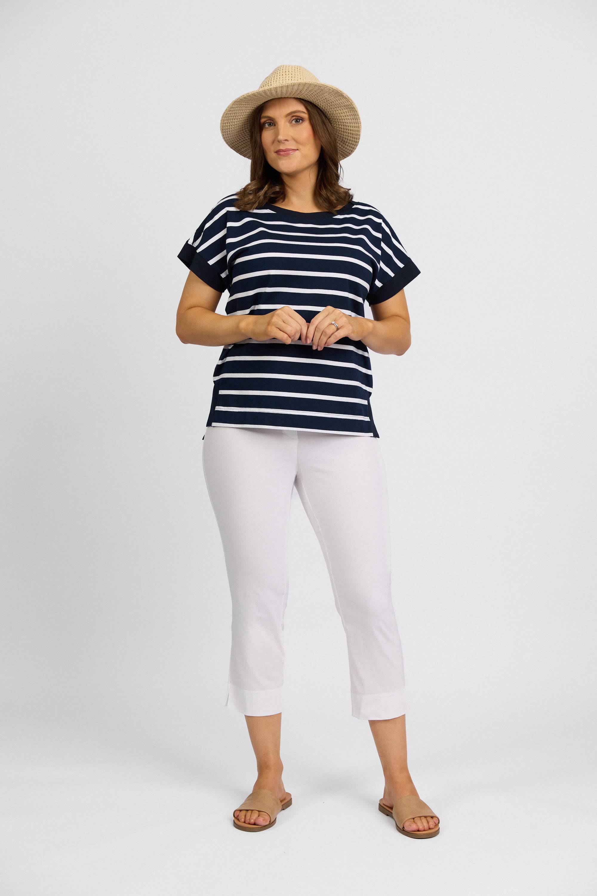 A woman wearing a straw hat, navy and white striped short-sleeve shirt, Vassalli Paris Crop Pant in White, and sandals stands against a plain white background.