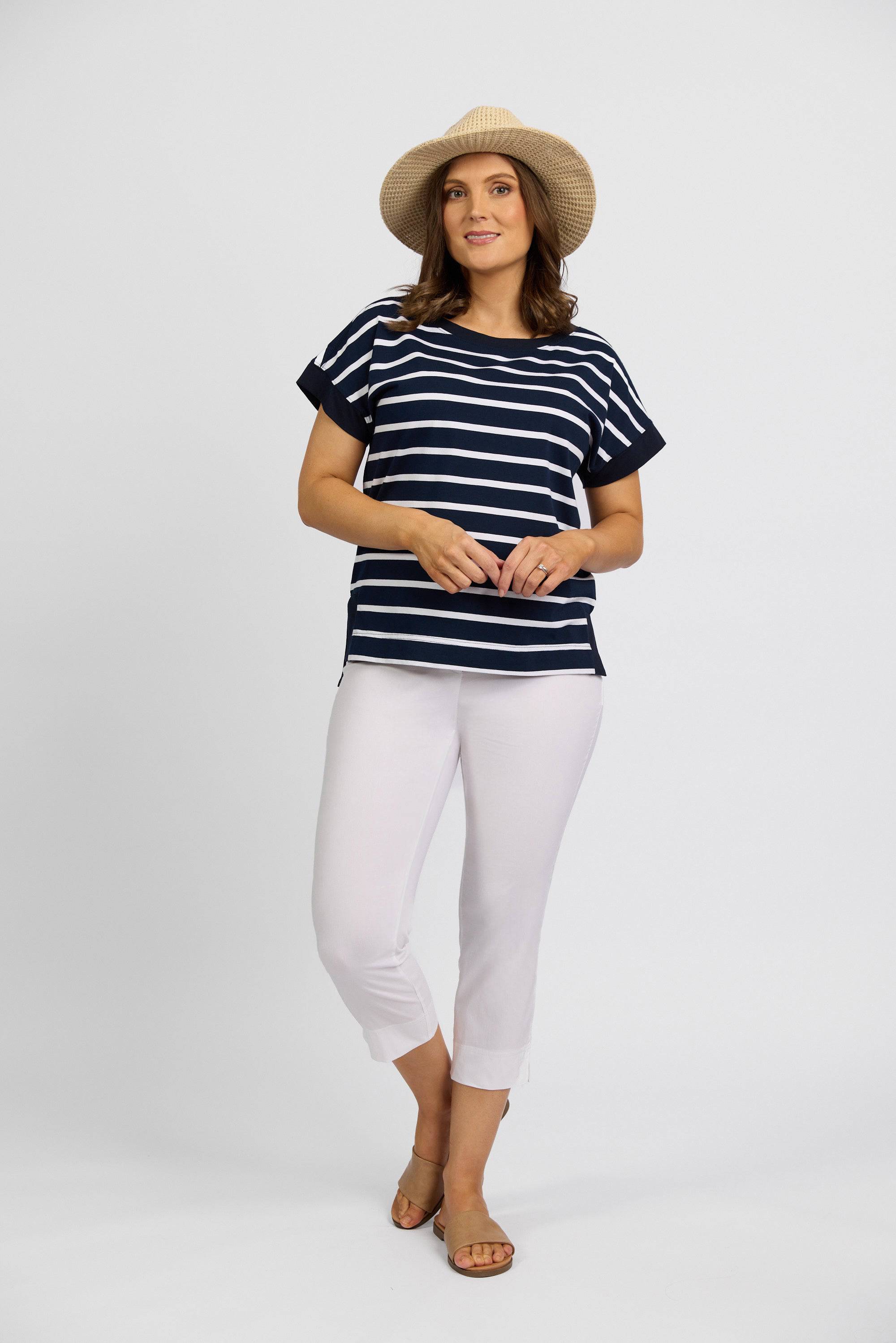 A woman wearing a straw hat, navy and white striped short-sleeve top, Vassalli Paris Crop Pant - White, and tan sandals stands against a plain light background.