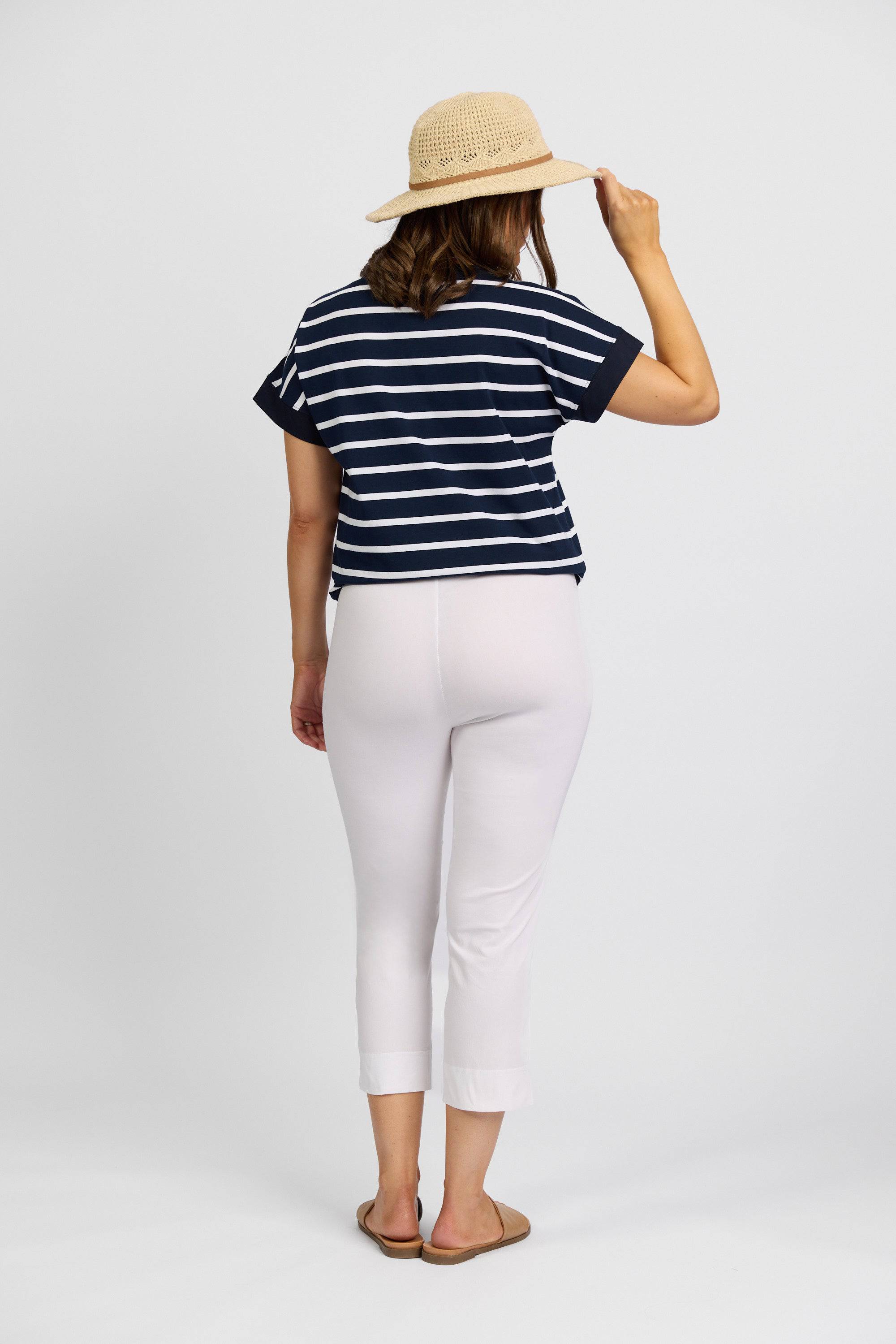 A woman faces away against a white background, wearing a straw hat, navy and white striped shirt, sandals, and Vassalli's Paris Crop Pant in White.