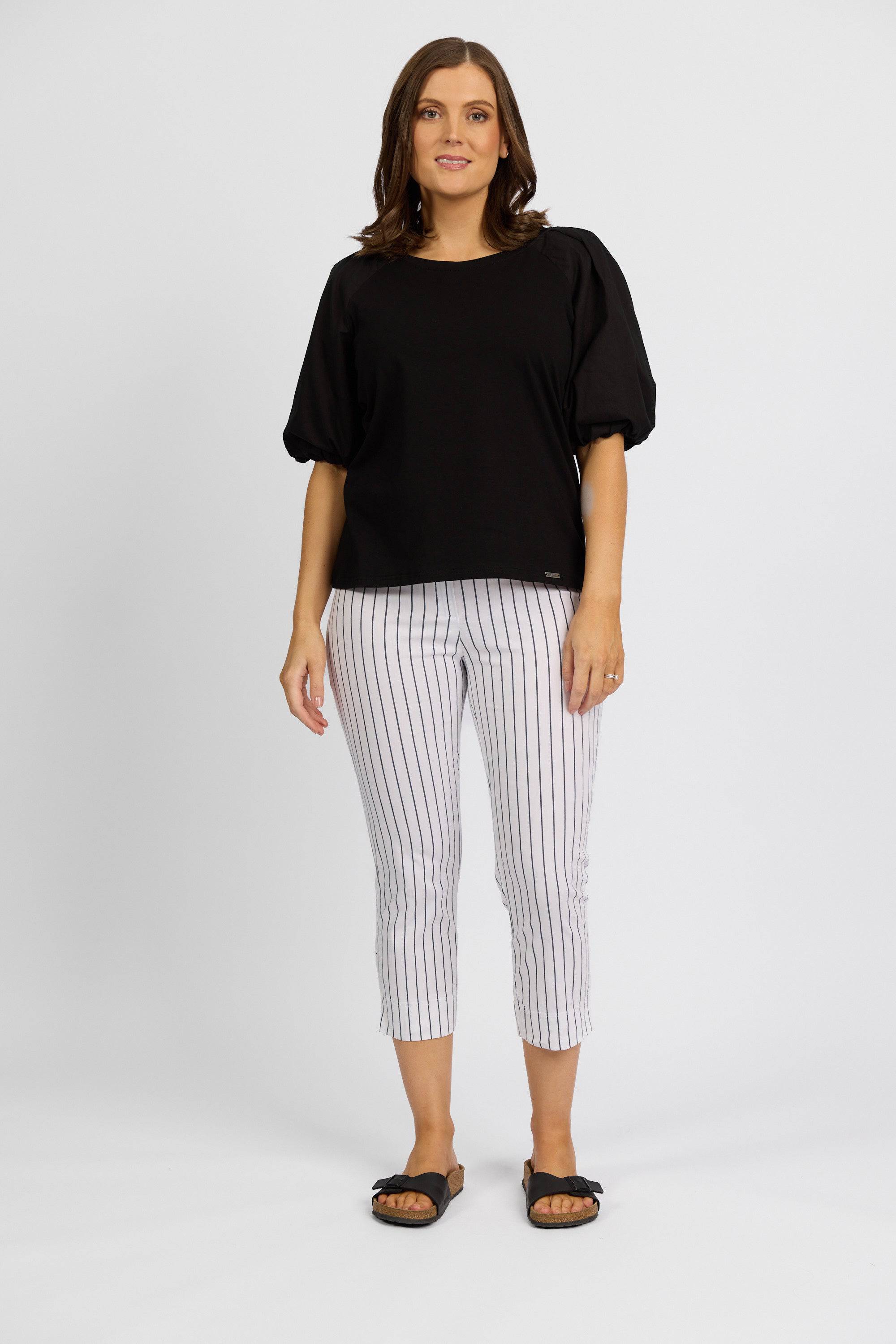 A woman with shoulder-length brown hair smiles against a plain background, wearing a black puff-sleeve top, black slide sandals, and Vassalli Paris Printed Crop Pant in white with stripes.