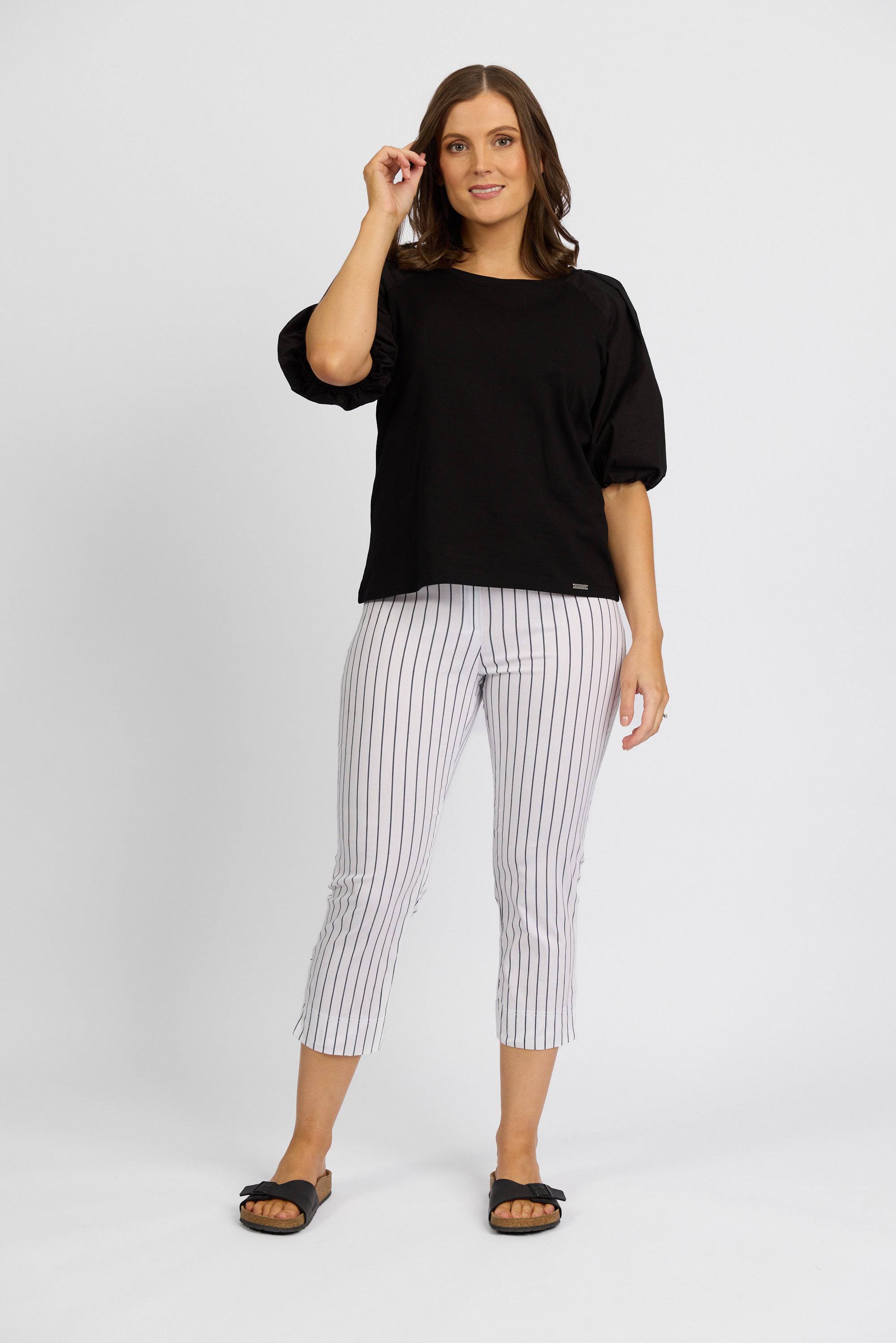 A woman with shoulder-length brown hair smiles softly, wearing a black top and Vassalli Paris Printed Crop Pant in white with black stripes, made in New Zealand. She stands against a white background.