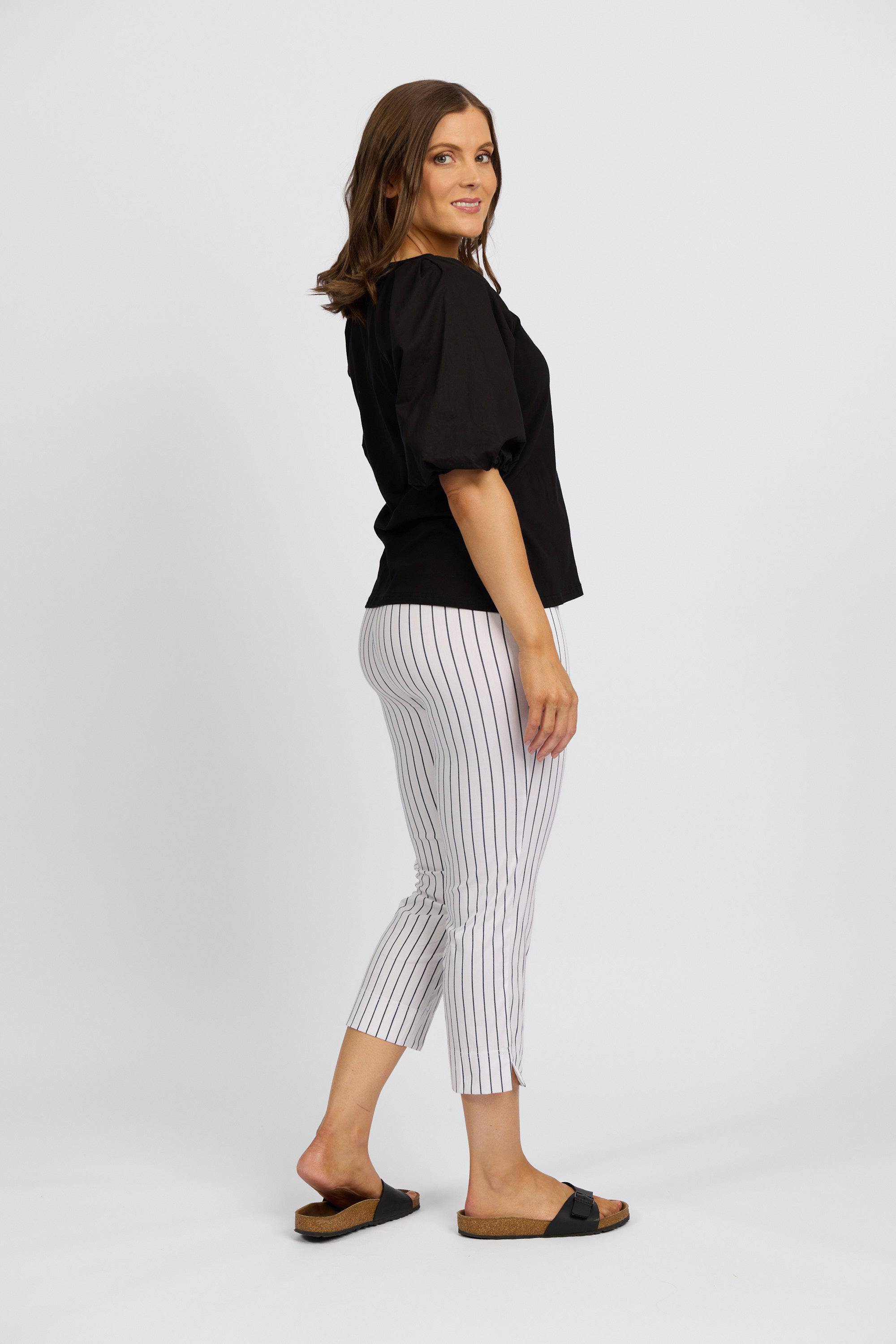 A woman smiles at the camera, standing sideways against a white background in a black puff-sleeve top, black slide sandals, and Vassalli's Paris Printed Crop Pant in white with vertical stripes.