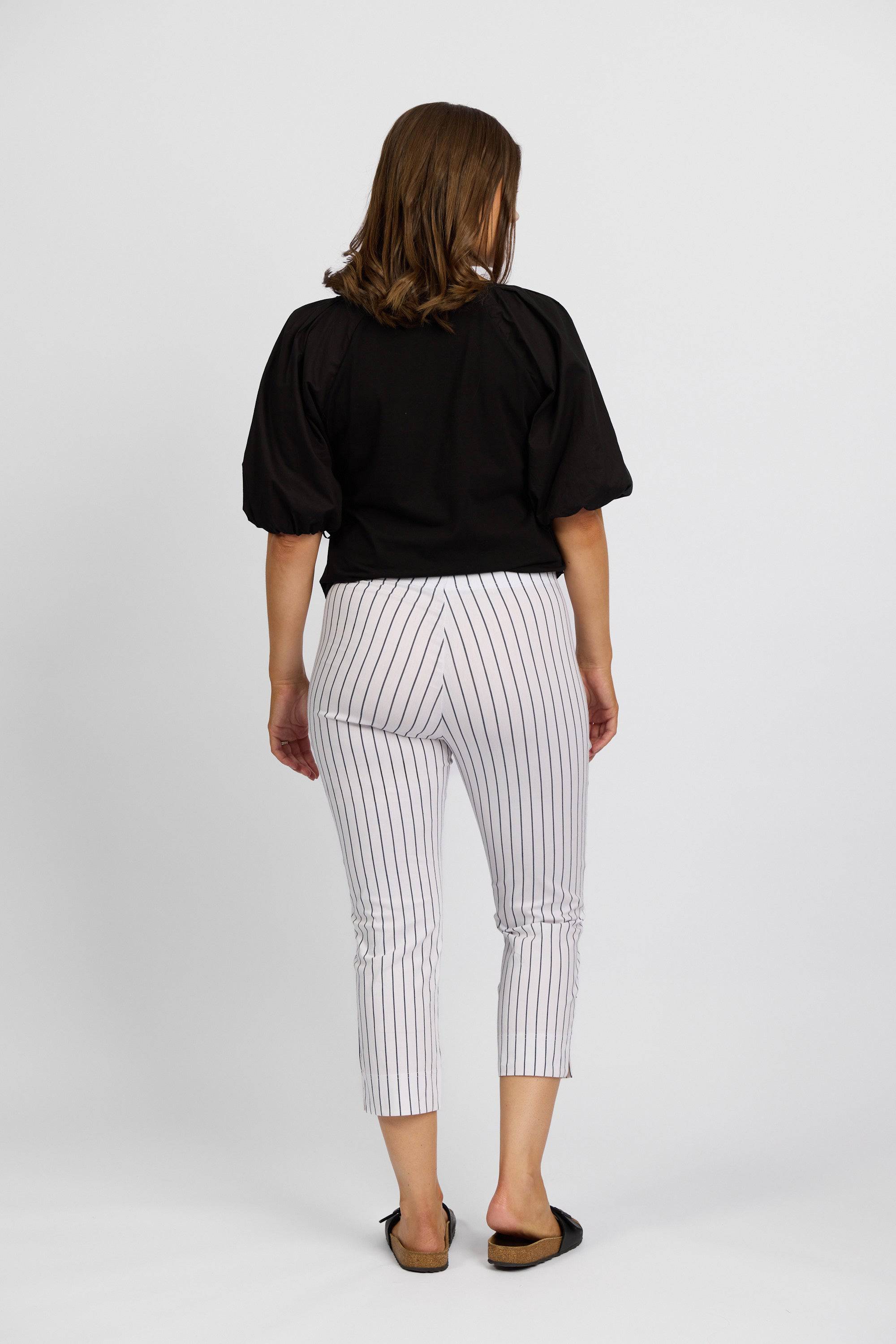 A woman faces away, wearing a black short-sleeve top and Vassalli Paris Printed Crop Pant in white with thin black stripes, made from stretch fabric, paired with black slip-on shoes, set against a plain white background.