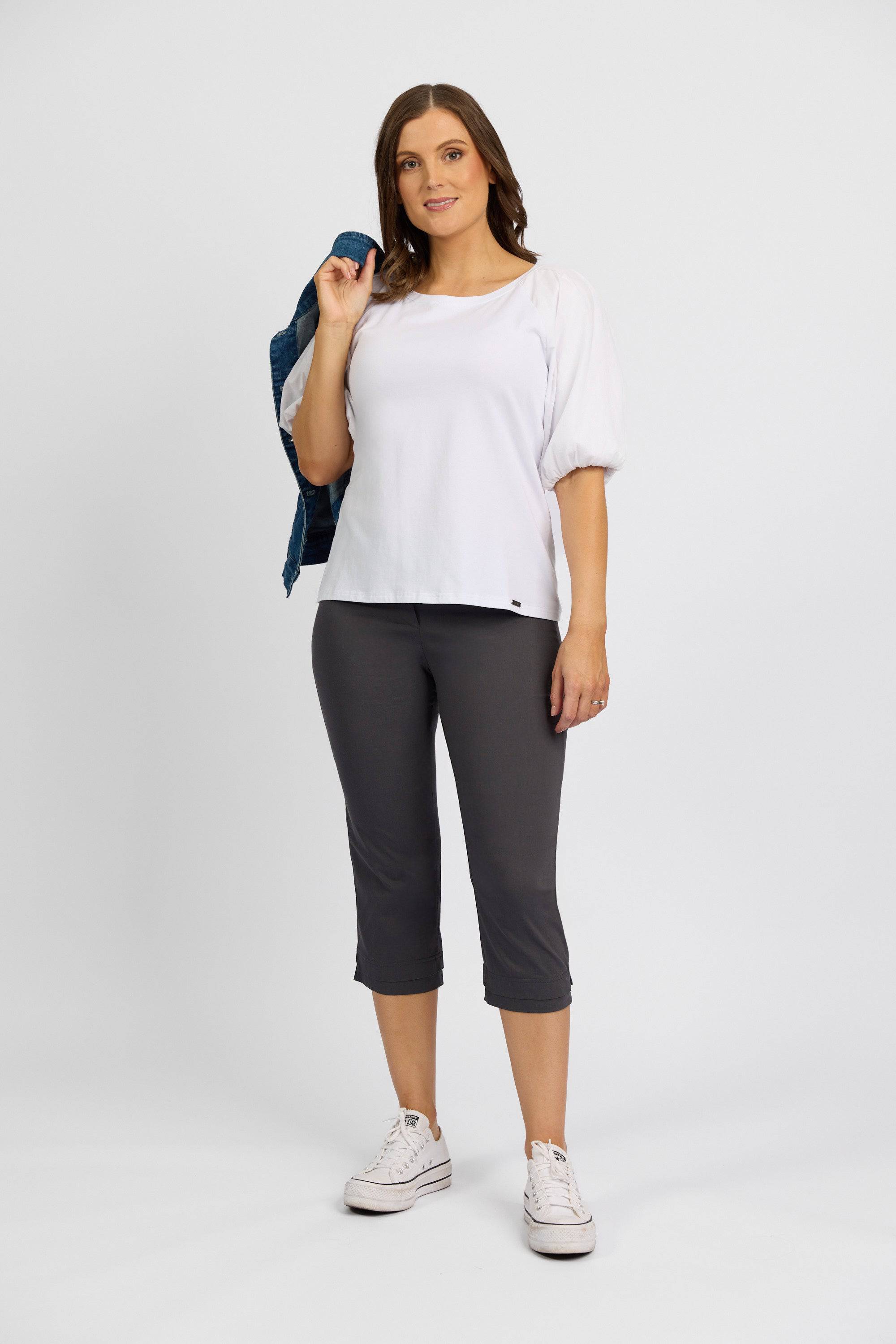 A woman stands against a plain background, wearing a white short-sleeve top, Vassalli Paris 3/4 Pant - Beluga, and white sneakers. She holds a denim jacket over her shoulder and smiles at the camera.