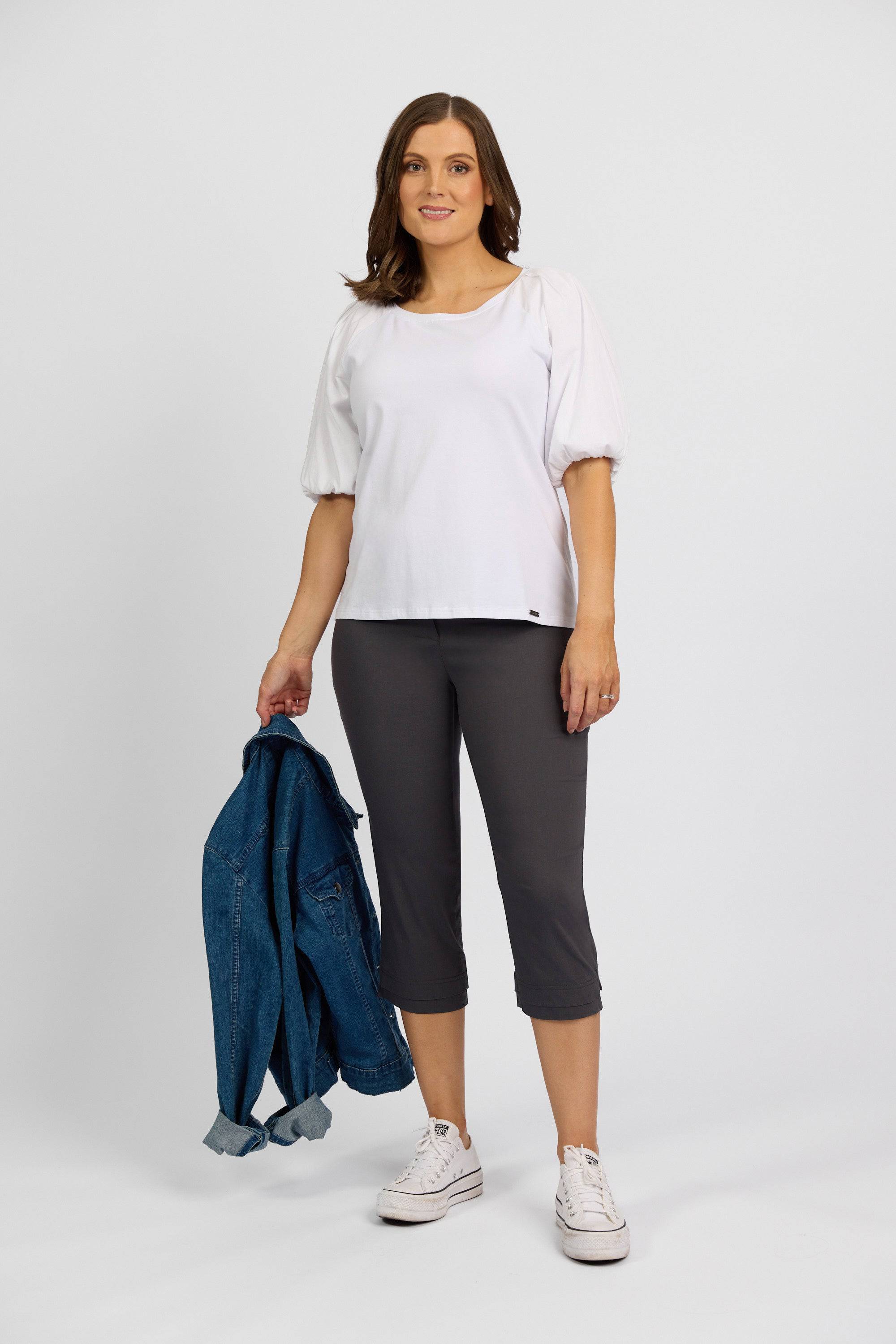 A woman smiles at the camera in a white blouse, white sneakers, and Vassalli Paris 3/4 Pant - Beluga. She holds a denim jacket with one hand against a plain background.