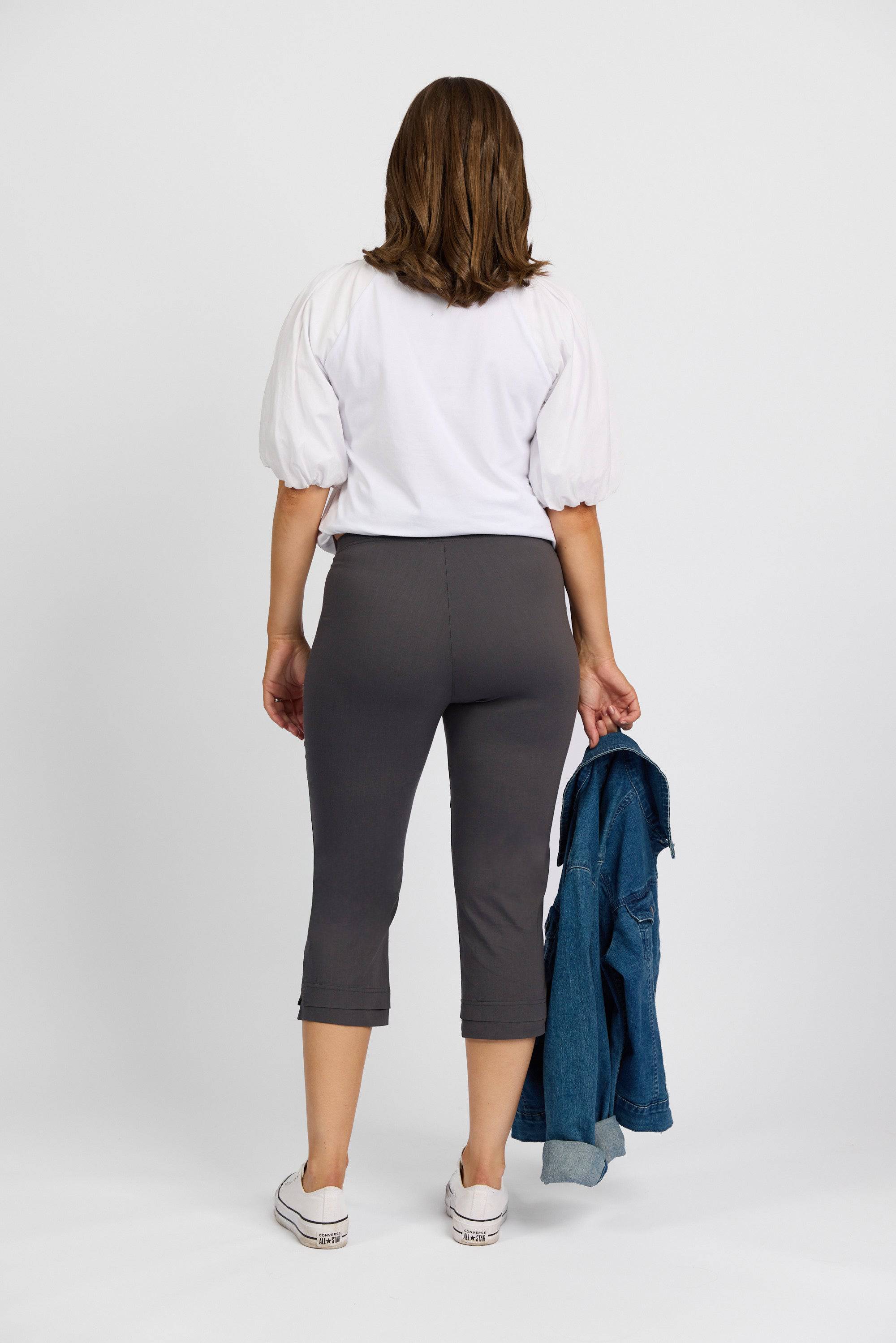 A woman faces away, wearing a white short-sleeve top, Vassalli Paris 3/4 Pant - Beluga with stretch and mid rise, and white sneakers. She holds a blue denim jacket in her left hand against a plain white background.