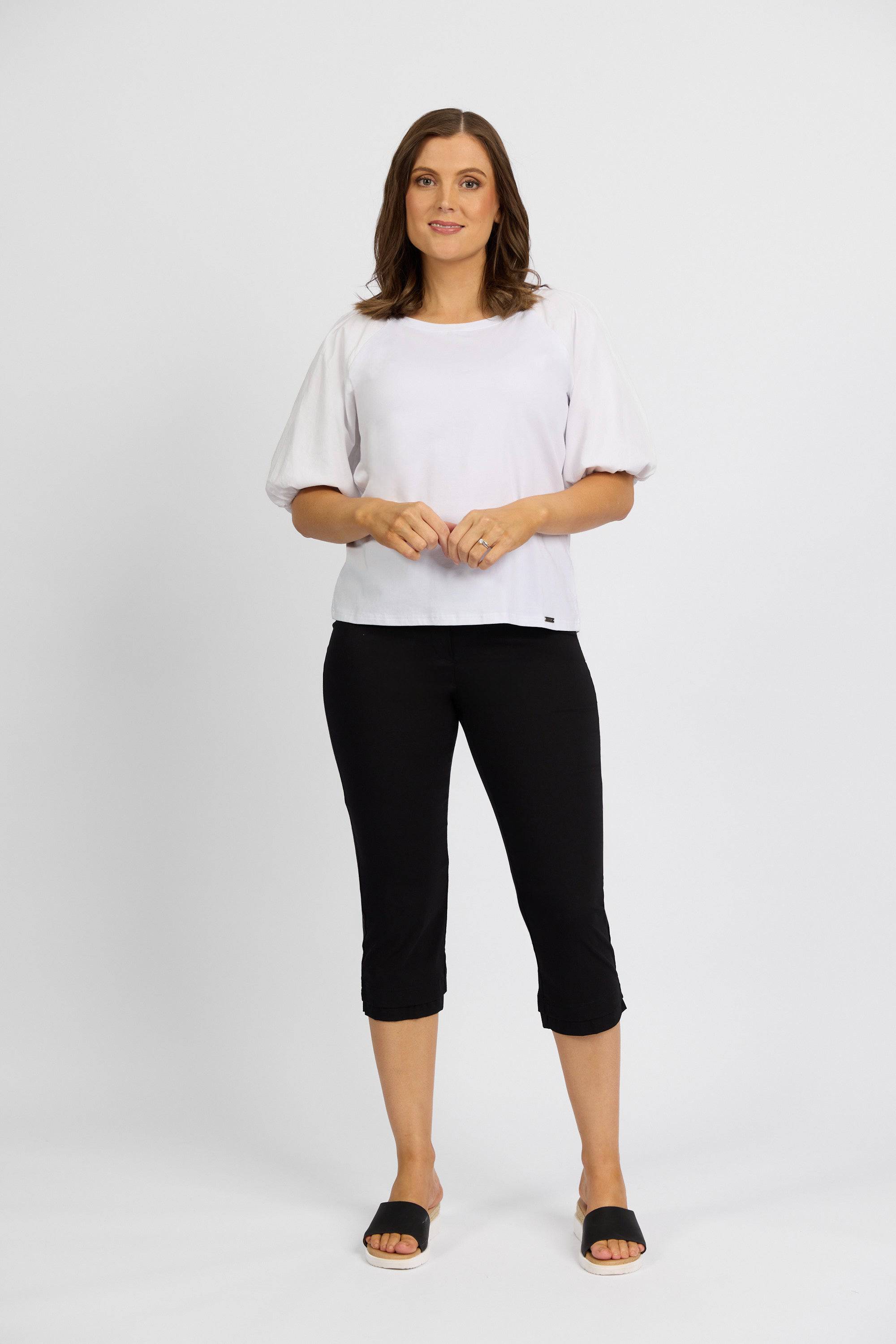 A woman stands against a white background in a loose white short-sleeve top and Vassalli Paris 3/4 Pant - Black, styled with black slide sandals. She smiles slightly, hands lightly clasped in front.