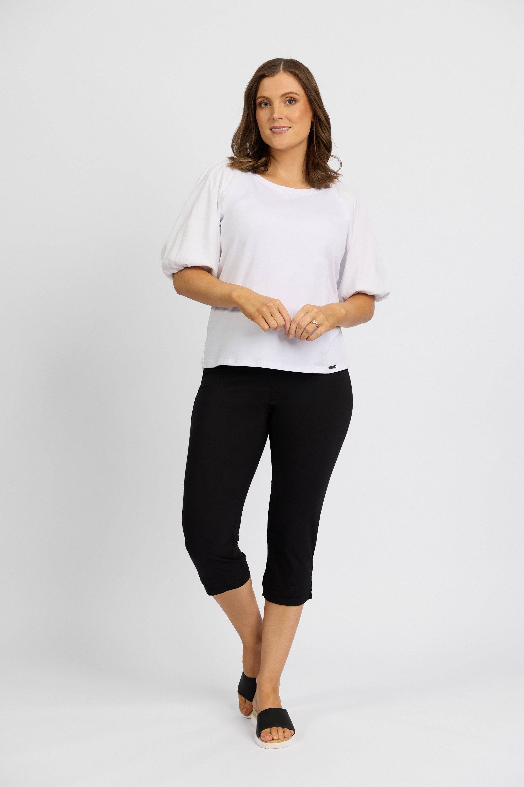 A woman stands against a plain light background, wearing the Vassalli Paris 3/4 Pant in black with a white short-sleeve top and black slide sandals. She smiles gently and clasps her hands in front of her.