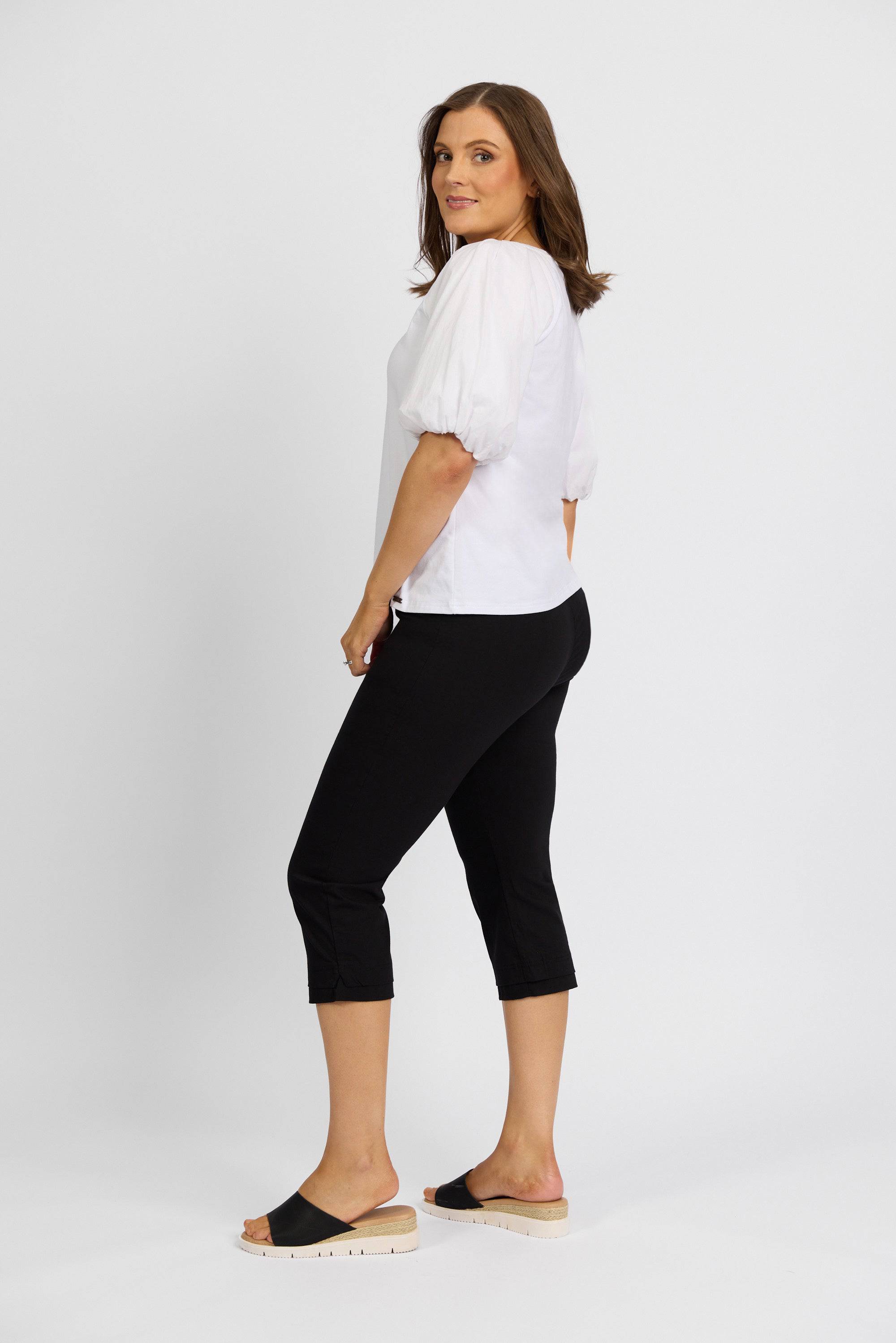 A woman stands sideways, gazing over her shoulder in a white blouse and Vassalli Paris 3/4 Pant - Black, paired with black open-toe slides. Her expression is neutral, set against a plain white background.
