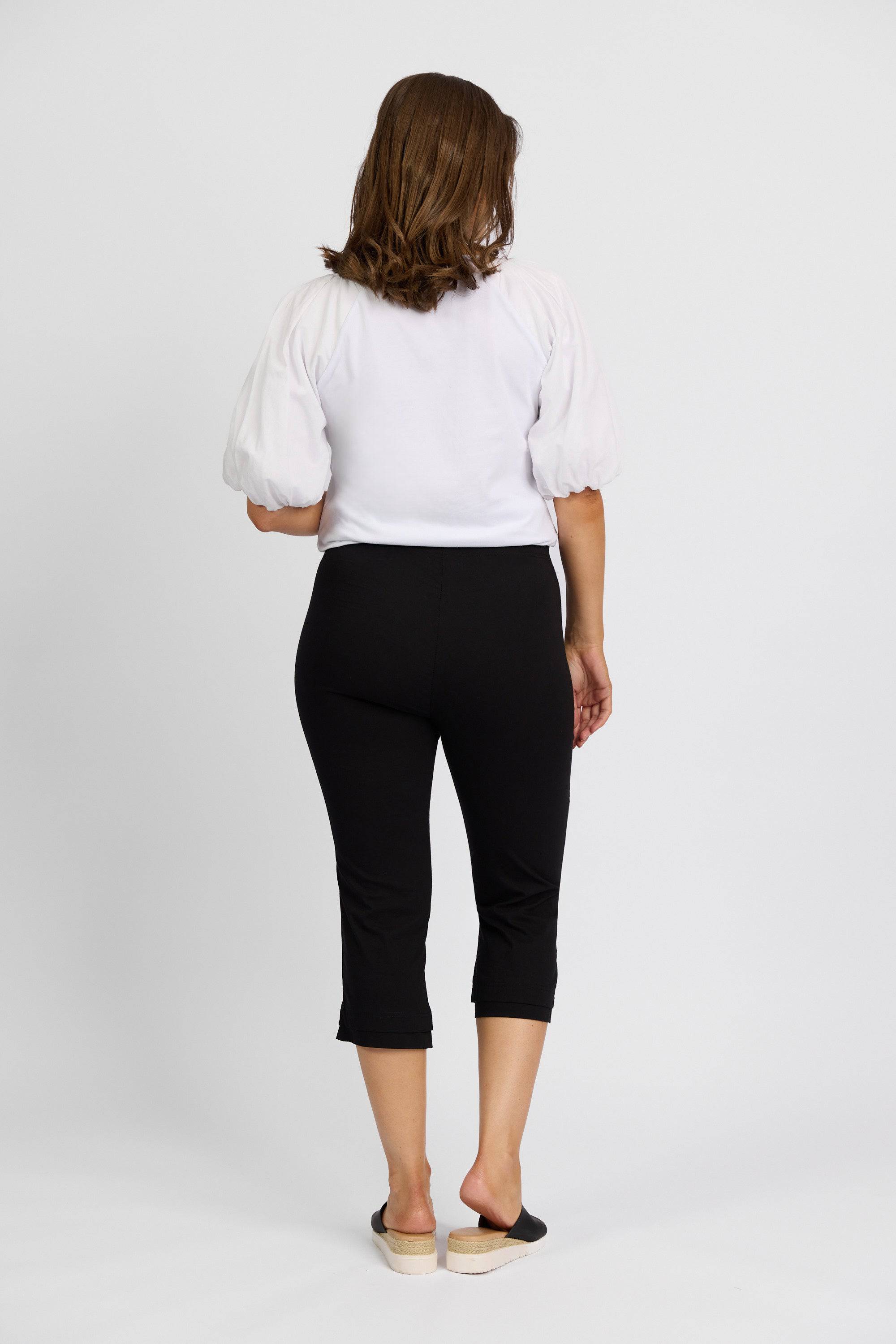A woman faces away, wearing a white puff-sleeve top and Vassalli Paris 3/4 Pant - Black, mid-rise cropped leggings in stretch fabric, with black slip-on shoes, against a plain white background.