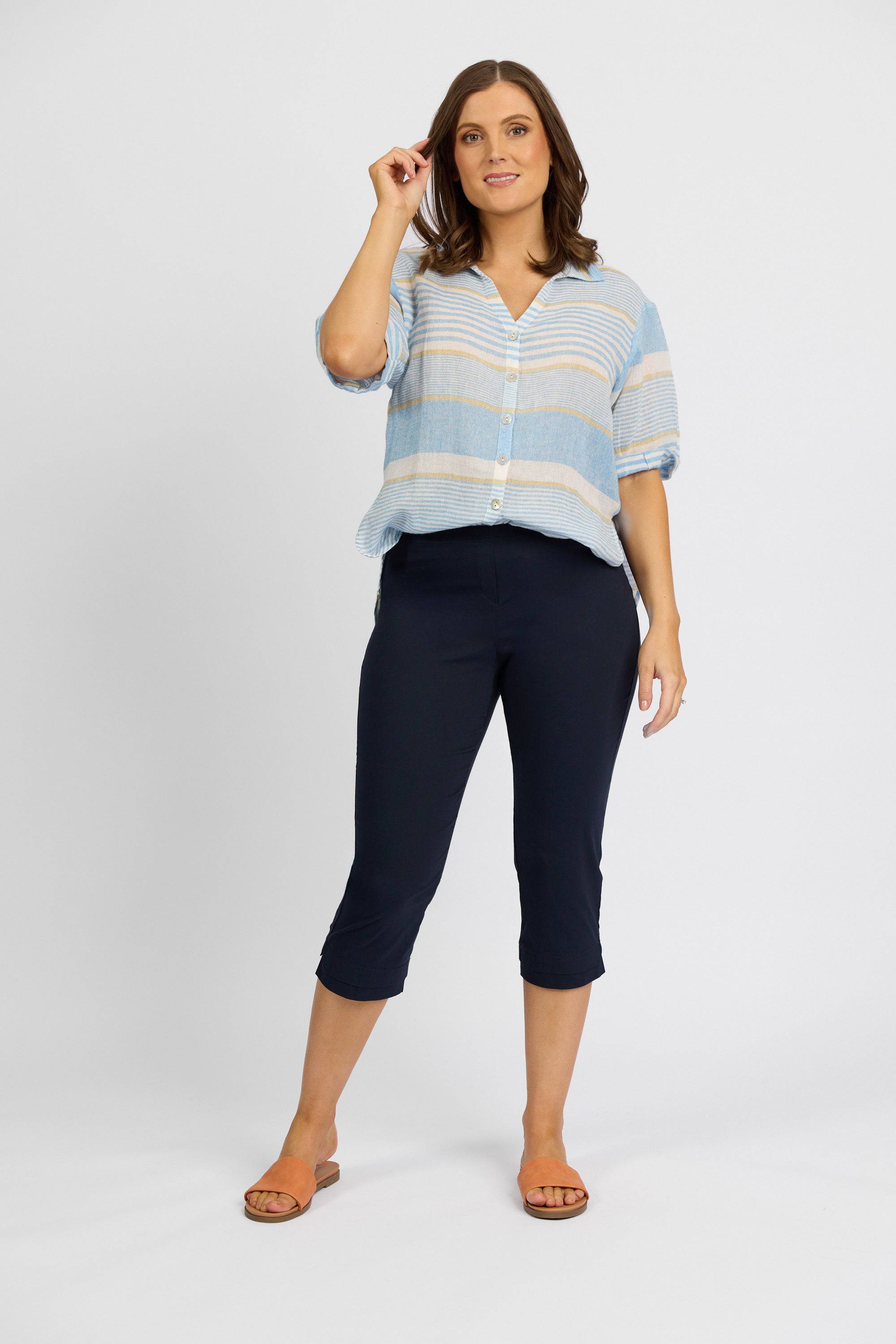 A woman poses against a white background, wearing Vassalli Paris 3/4 Pant - Inka with a blue and beige striped top and brown sandals, smiling and touching her hair—a chic look from the Paris Collection.