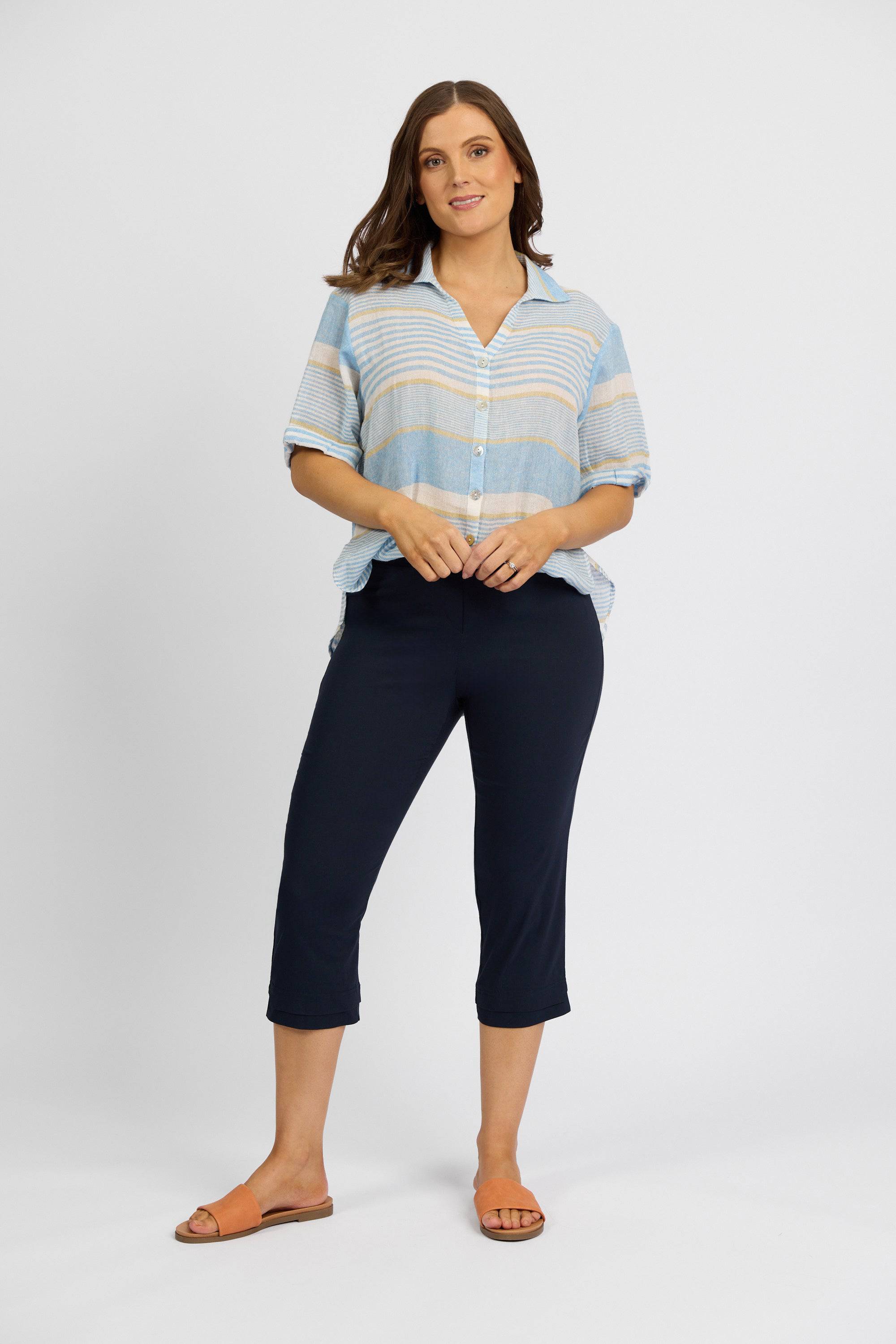 A woman stands against a white background, smiling slightly, wearing a light blue and beige striped button-up shirt and Vassalli Paris 3/4 Pant - Inka in navy stretch fabric, paired with orange flat sandals. One hand rests on her shirt.