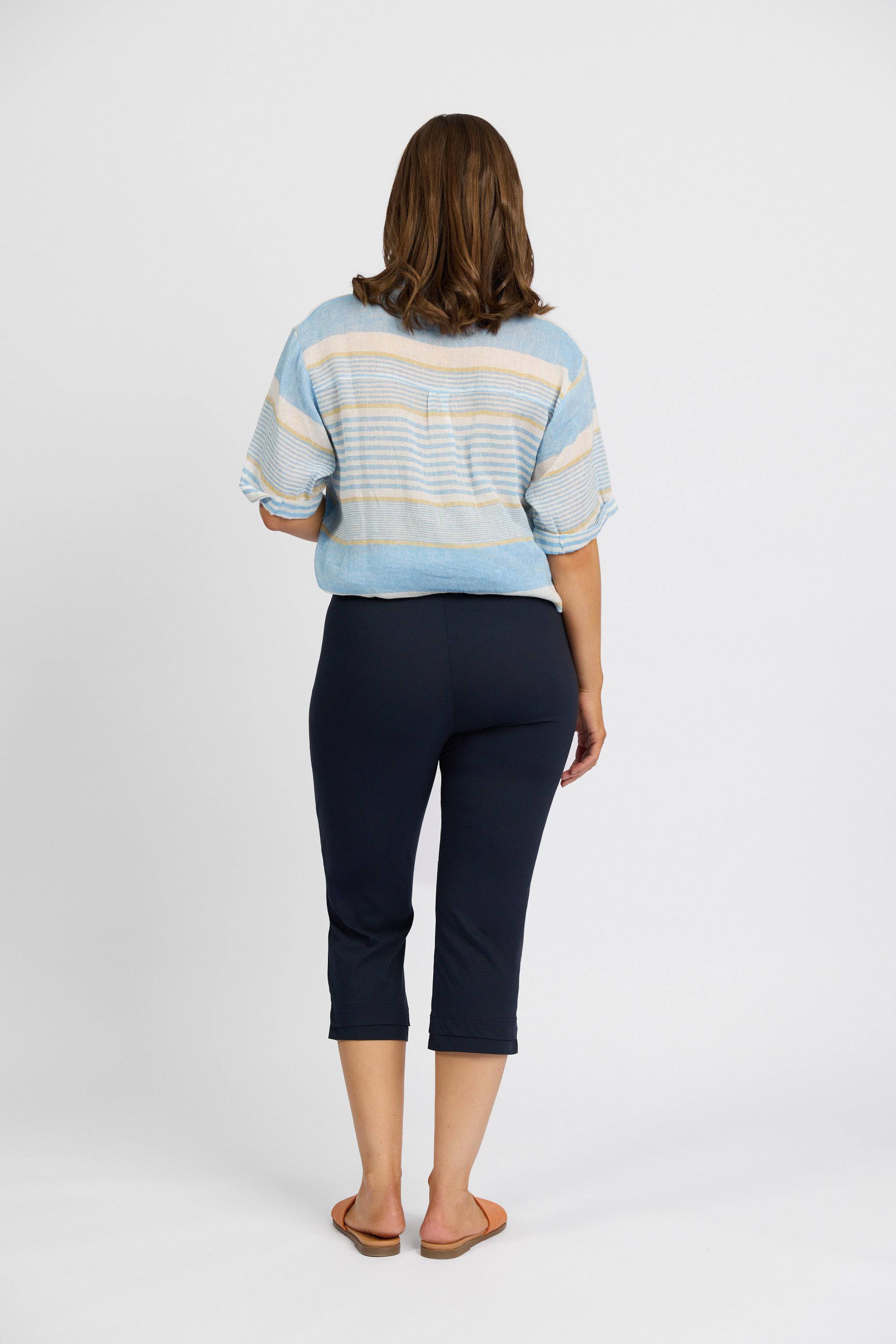 A woman stands facing away, wearing a light blue striped top, the Vassalli Paris 3/4 Pant - Inka in navy stretch fabric, and brown sandals against a plain white background.