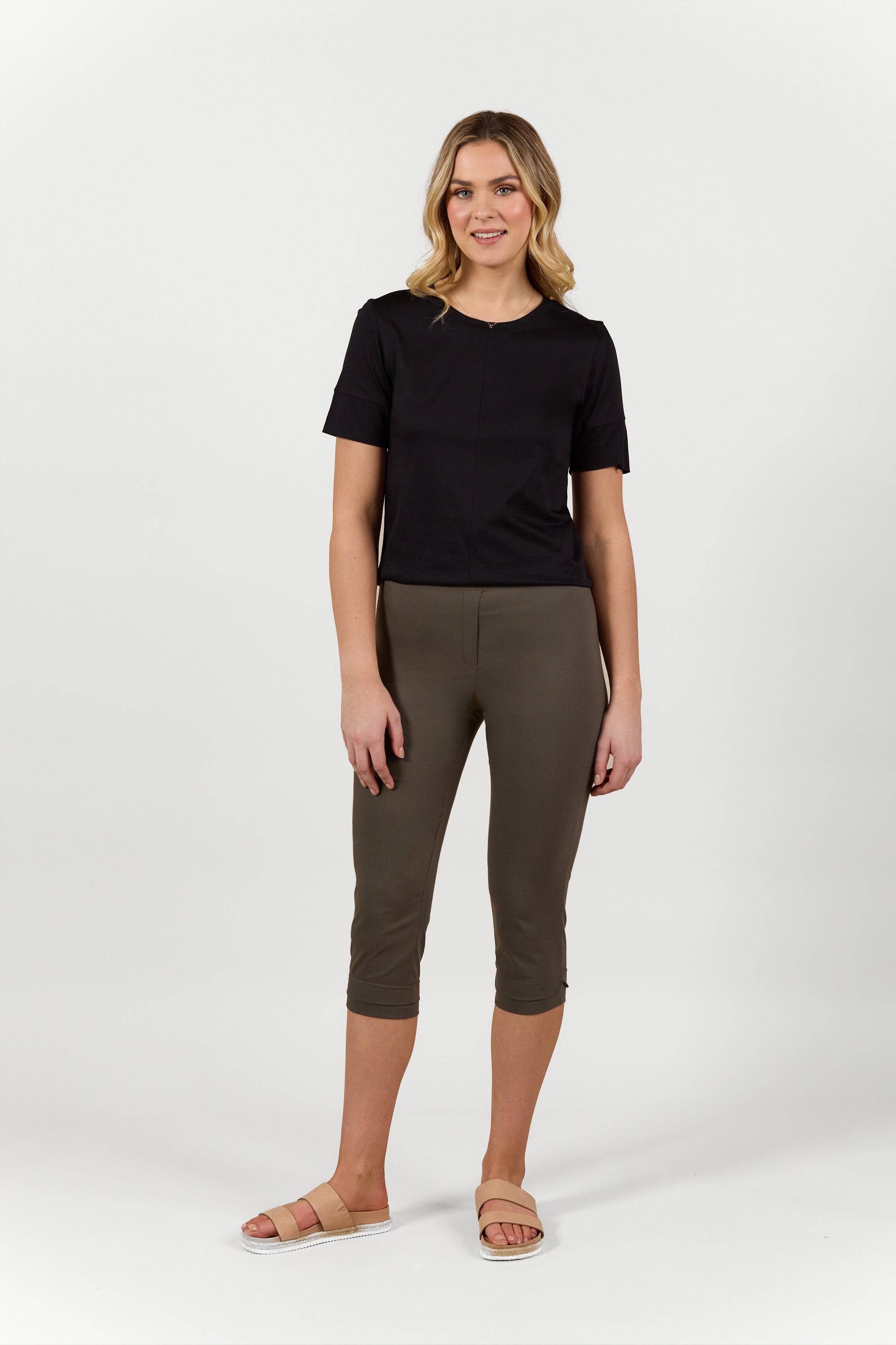 A woman smiles at the camera, wearing a black short-sleeve top and Vassalli Paris 3/4 Pant in khaki, styled with beige sandals against a plain light background.