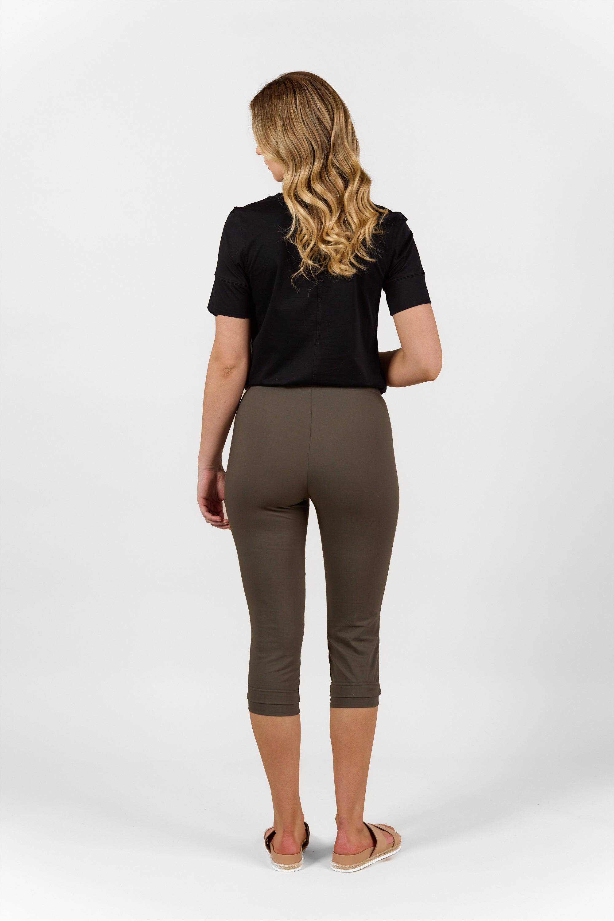 A woman with wavy blonde hair, in a black short-sleeve shirt and Vassalli Paris 3/4 Pant - Khaki, stands in sandals facing away against a plain white background.