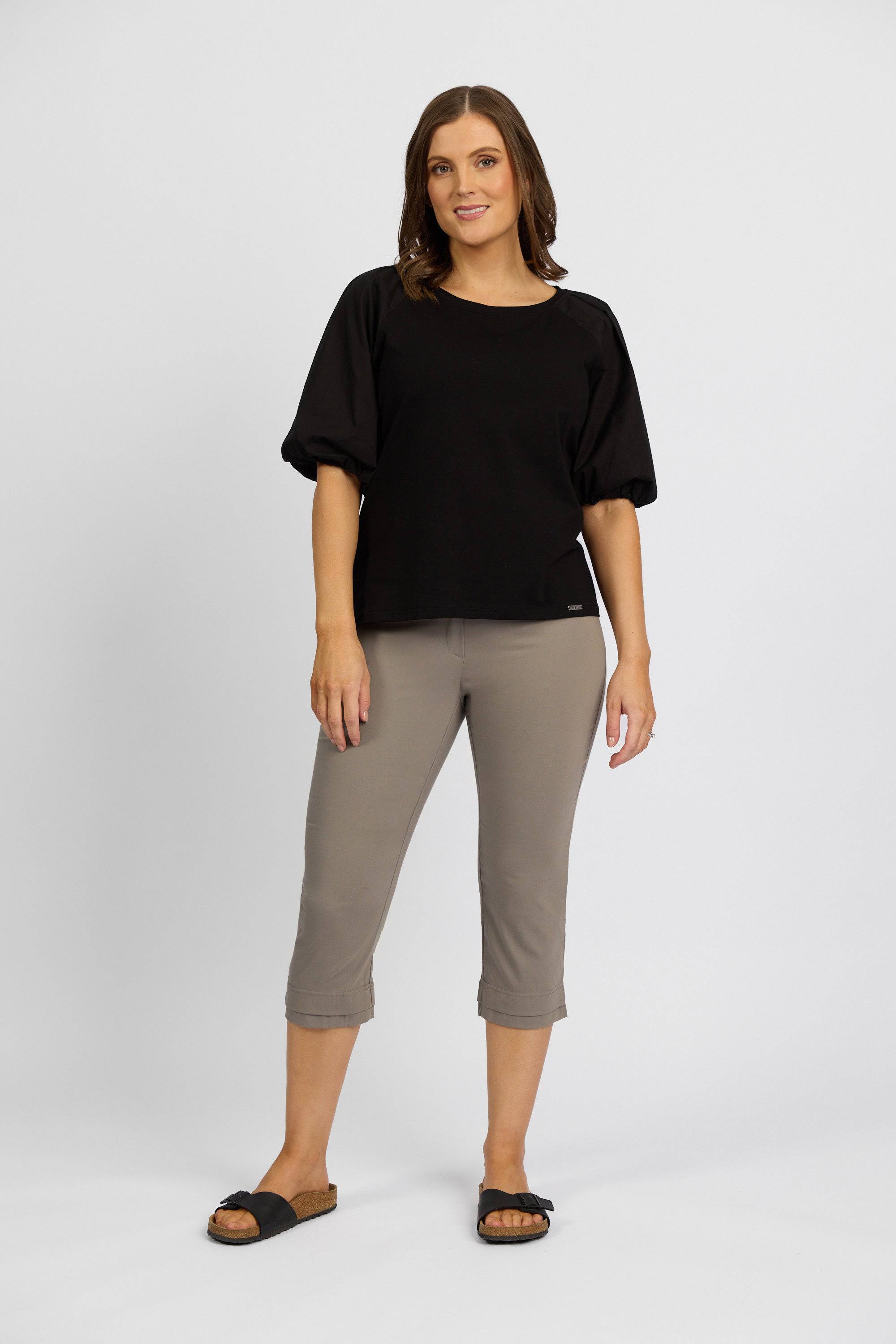 A woman with shoulder-length brown hair smiles slightly, wearing a black short-sleeve top, Vassalli Paris 3/4 Pant in Latte, and black slide sandals against a plain background.