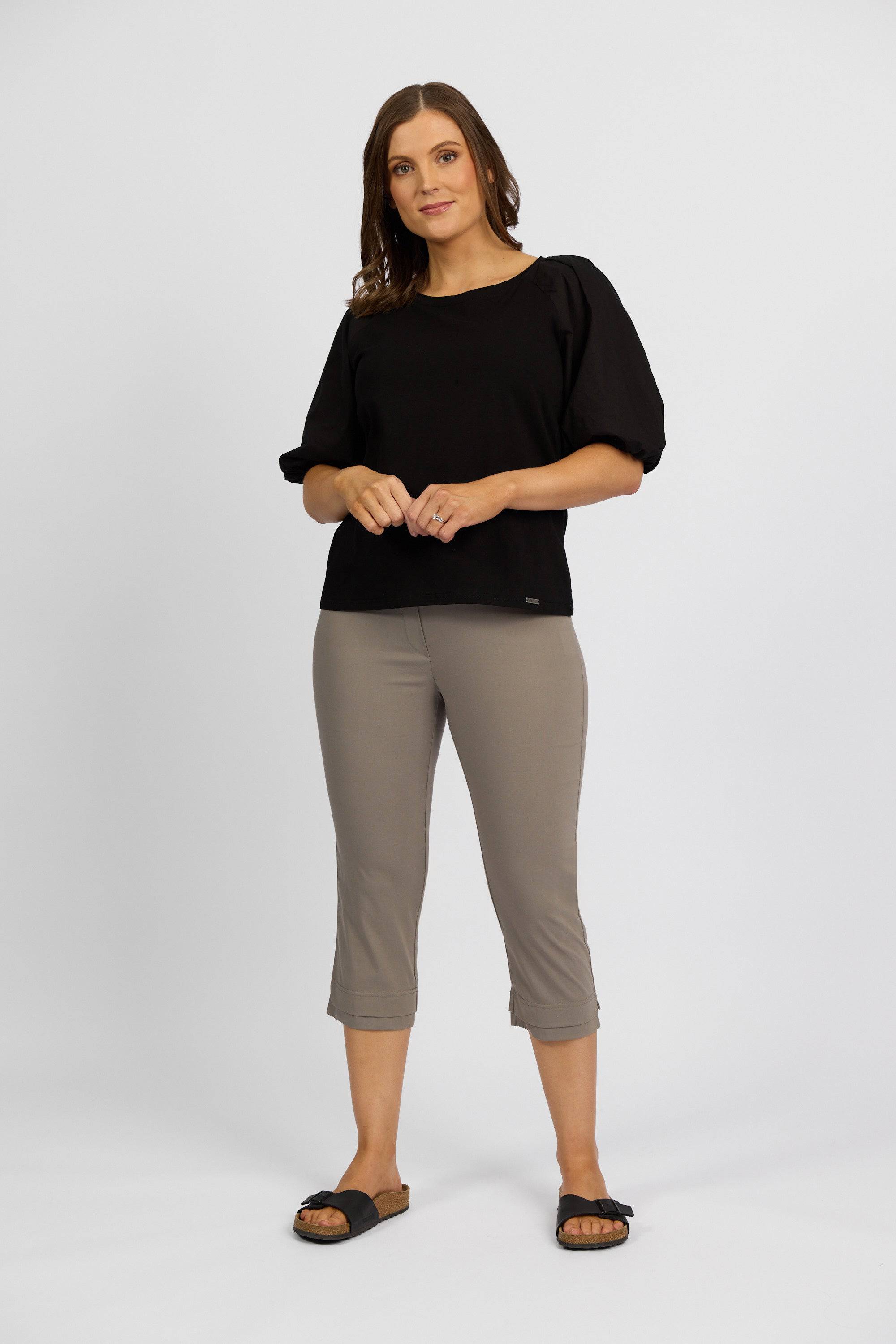 A woman stands against a plain background wearing a black top, Vassalli Paris 3/4 Pant in Latte crafted from stretch fabric, and black sandals. She faces forward with hands gently clasped, embodying effortless Paris Collection style.