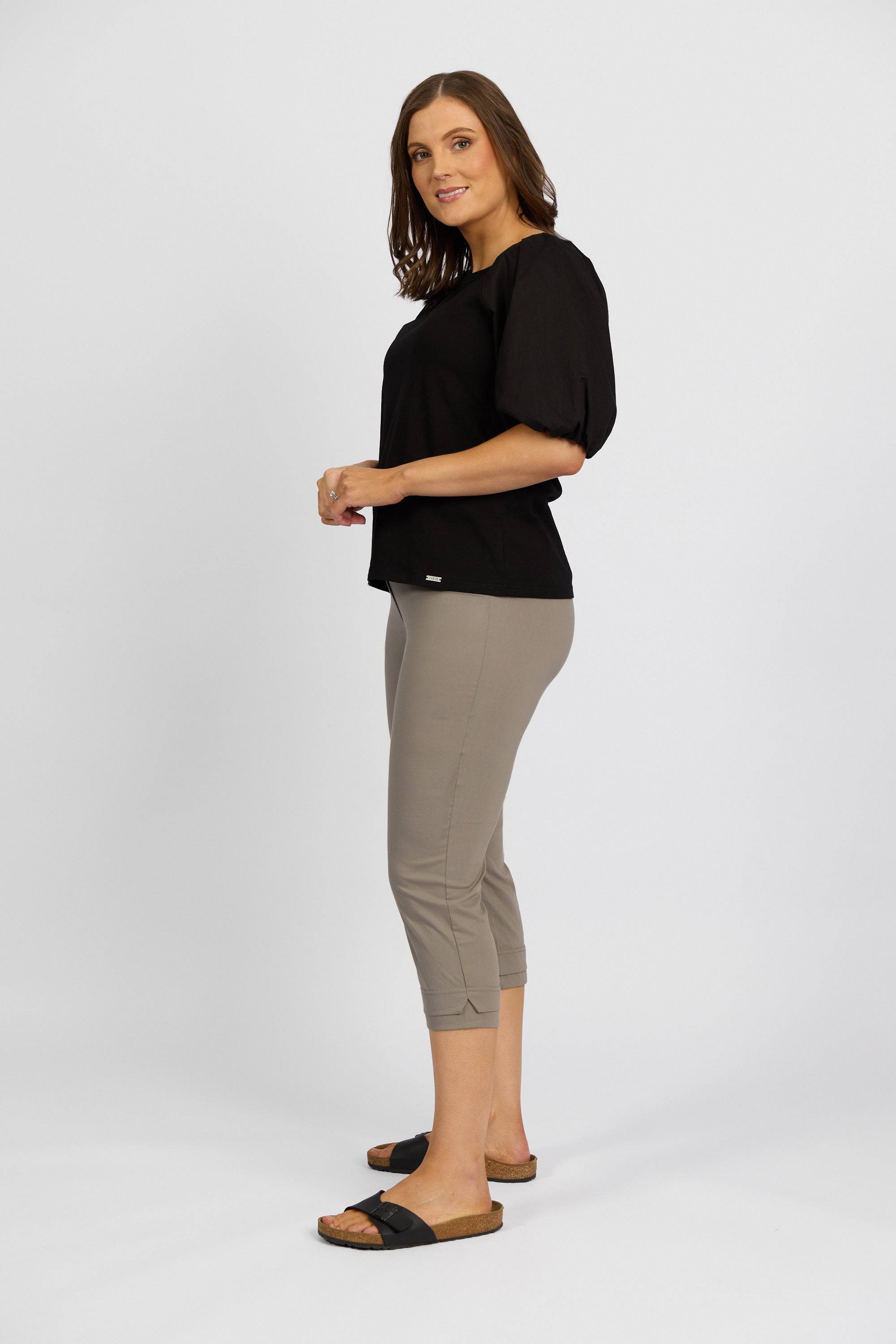 A woman with shoulder-length brown hair smiles gently while wearing a black short-sleeve top, Vassalli Paris 3/4 Pant - Latte, and black slide sandals, standing sideways against a plain white background.