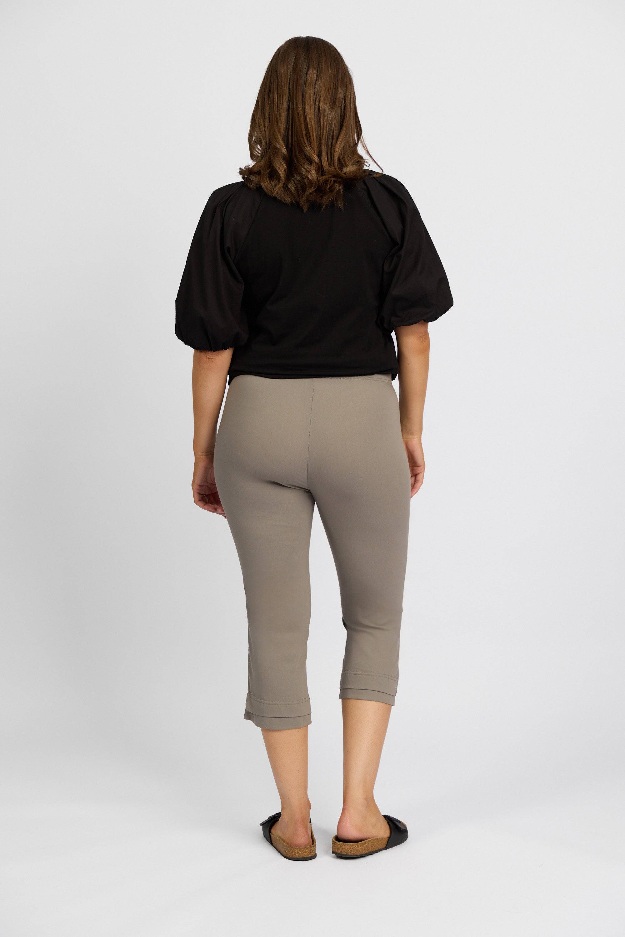 A woman wearing a black short-sleeved top, Vassalli Paris 3/4 Pant in Latte, and black slip-on shoes stands facing away from the camera against a white background.