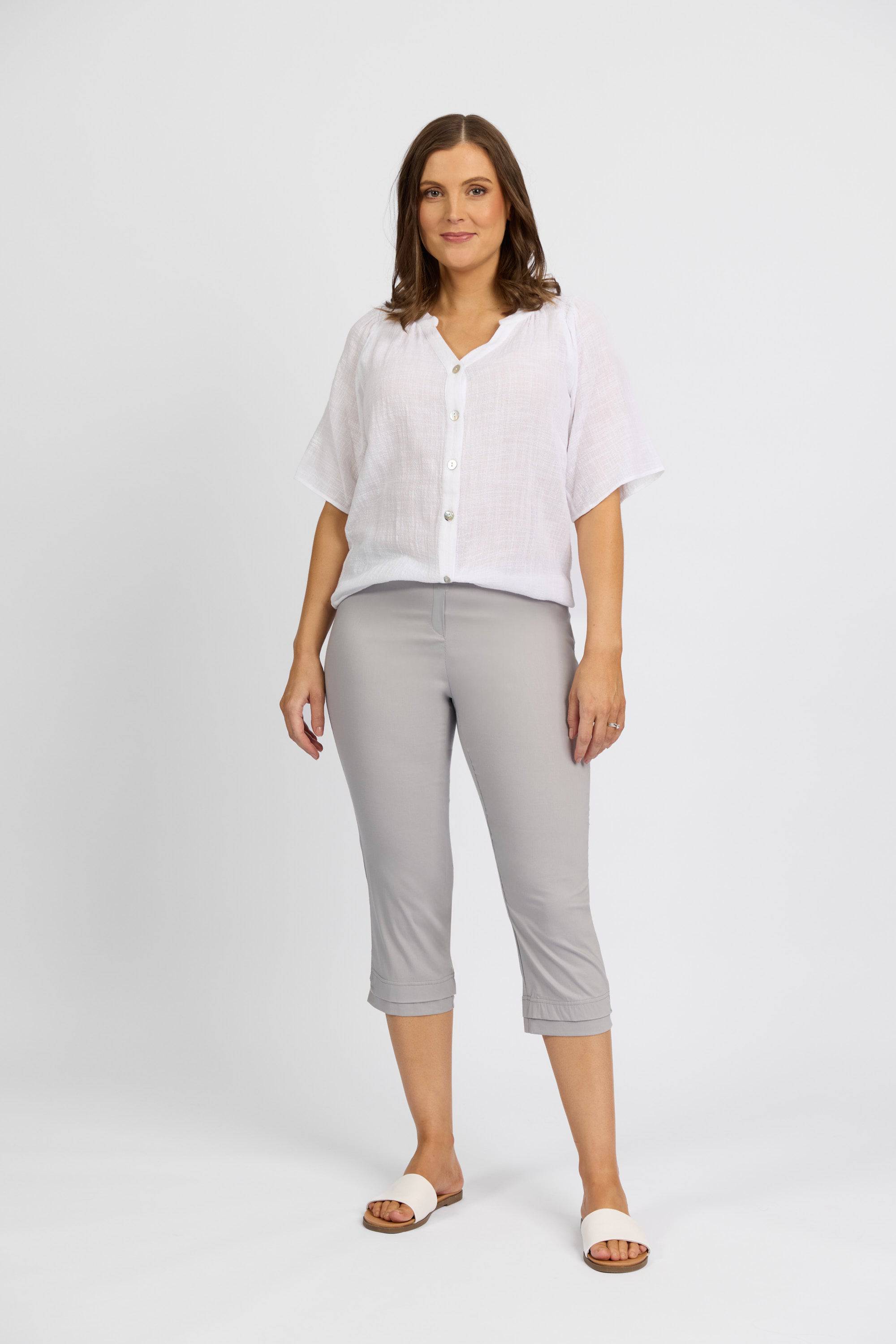 A woman with shoulder-length brown hair stands against a white background, wearing a loose white blouse and Vassalli Paris 3/4 Pant - Silver, exuding a relaxed, confident vibe.