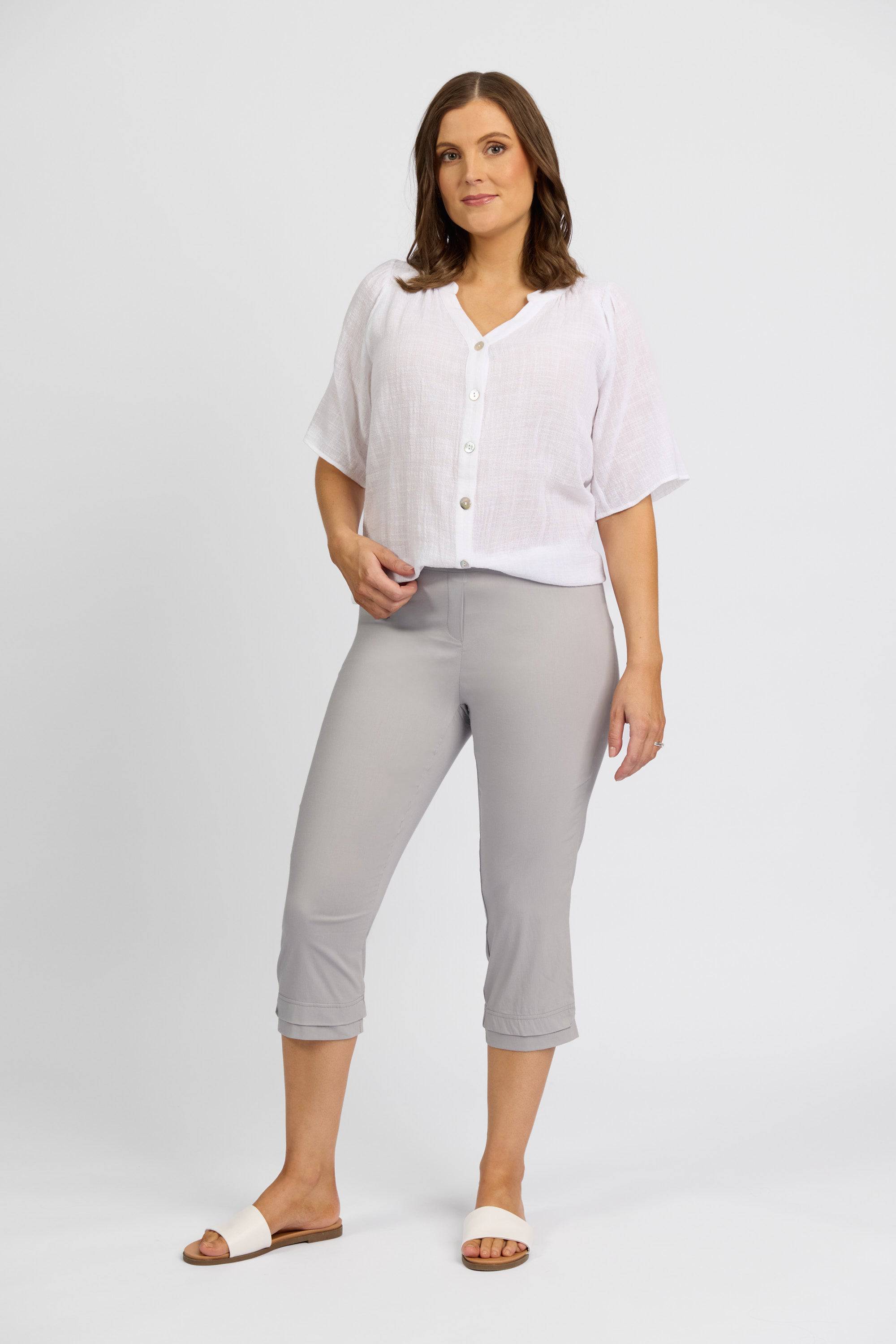 A woman with shoulder-length brown hair stands against a white background wearing a white short-sleeve top, Vassalli Paris 3/4 Pant in Silver, and white slide sandals. She has a neutral expression.
