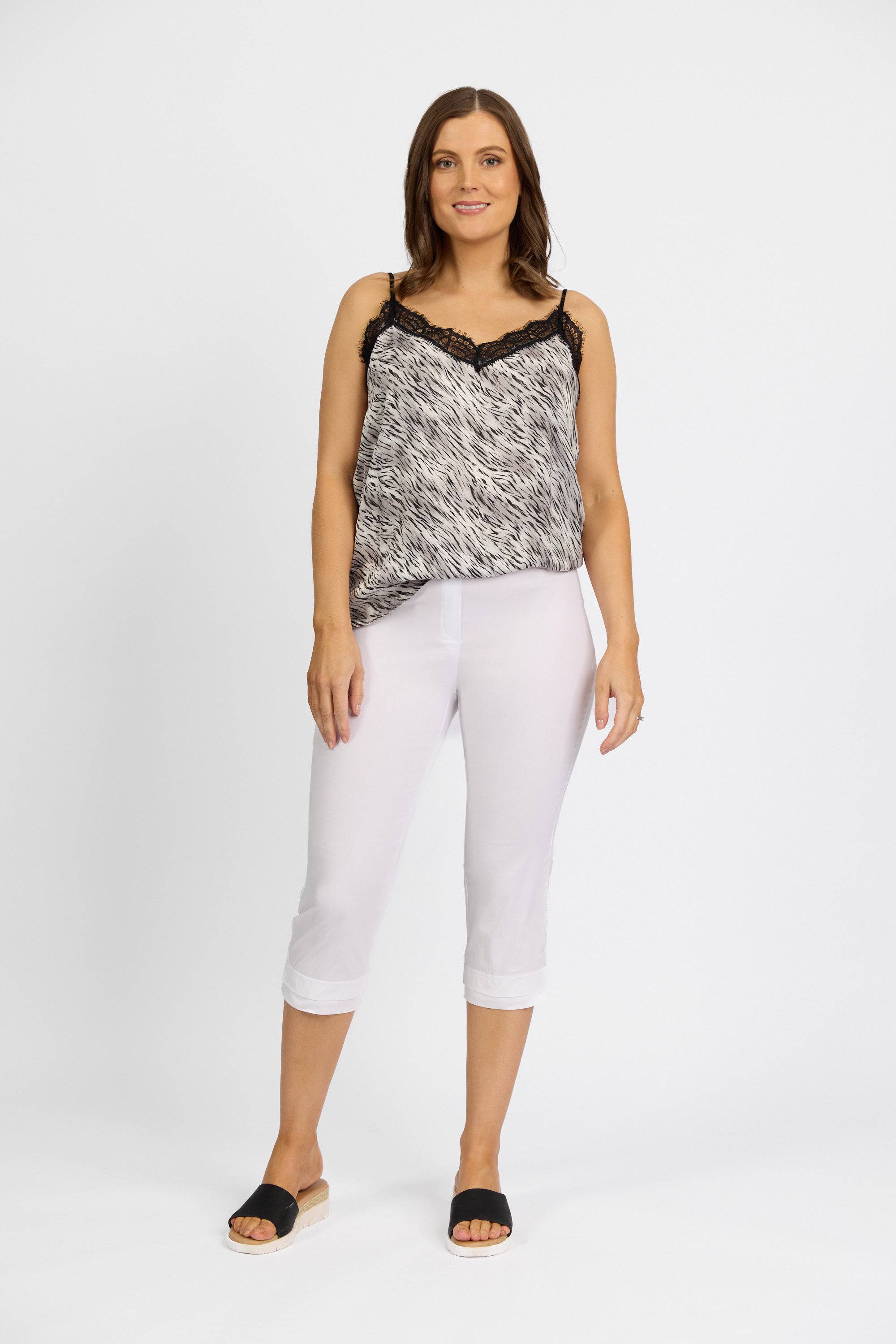 A woman with shoulder-length brown hair stands against a white background, smiling slightly. She wears a black and white patterned sleeveless top, Vassalli Paris 3/4 Pant in white, and black open-toe sandals.