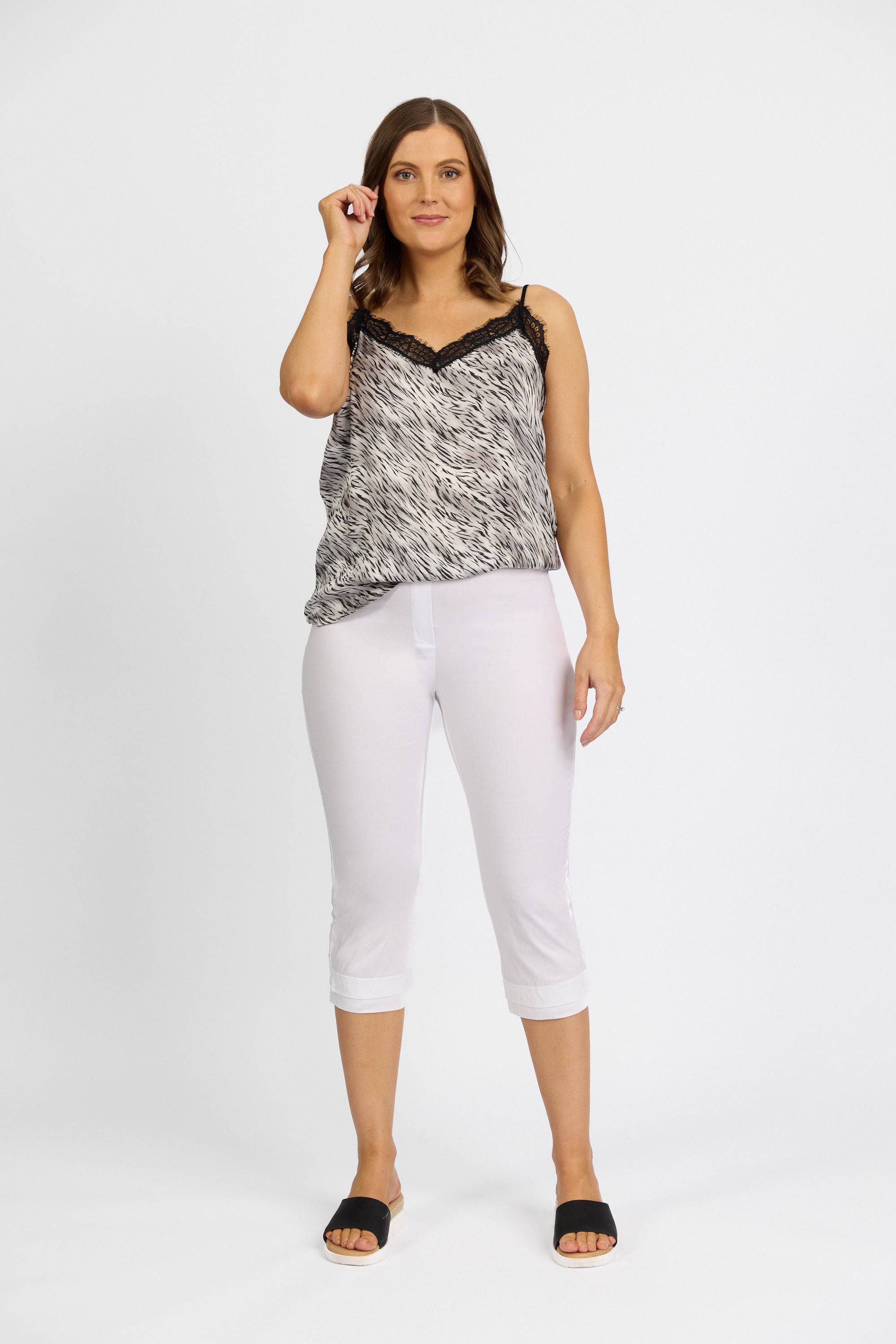 A woman stands against a white background, smiling and holding one hand near her face. She wears a black and white patterned sleeveless top, Vassalli Paris 3/4 Pant in white, and black slide sandals.