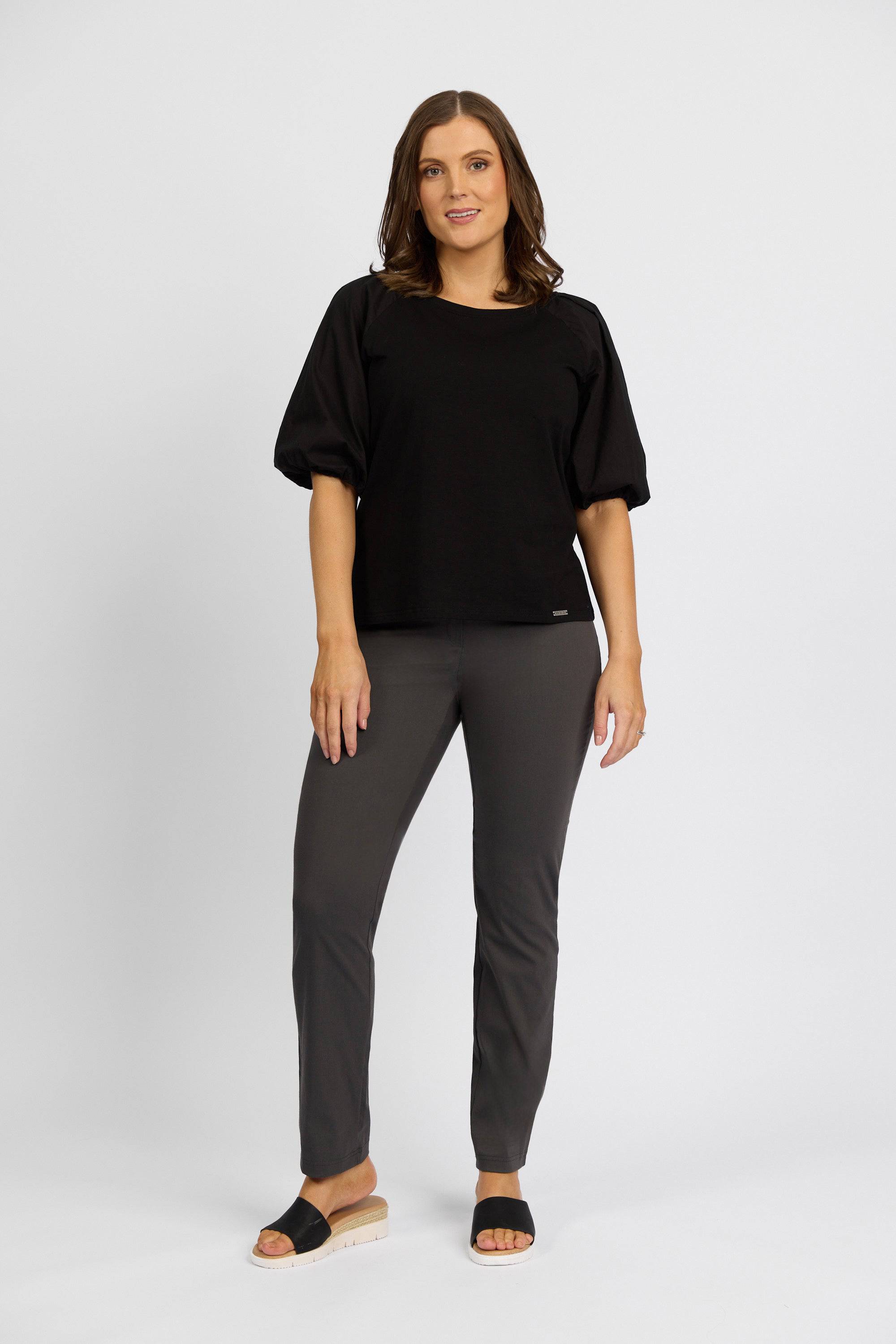 A woman with shoulder-length brown hair smiles softly as she models Vassalli’s Paris Full Length Pant – Beluga, styled with a black short-sleeve top and black platform open-toe sandals against a plain white background.