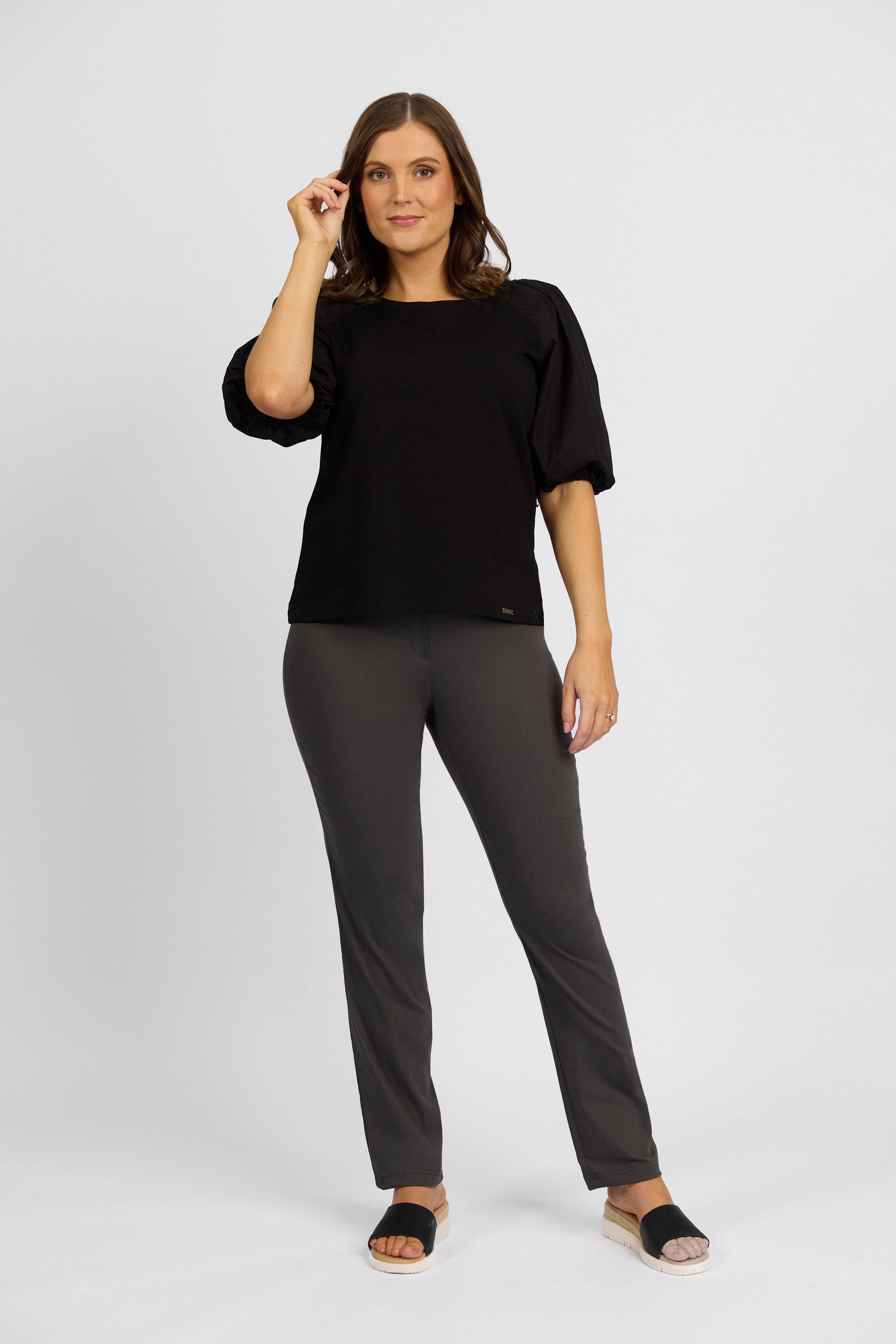 A woman with shoulder-length brown hair stands against a plain white background, wearing a black top and Vassalli’s Paris Full Length Pant in Beluga. She looks forward, one hand raised near her face.