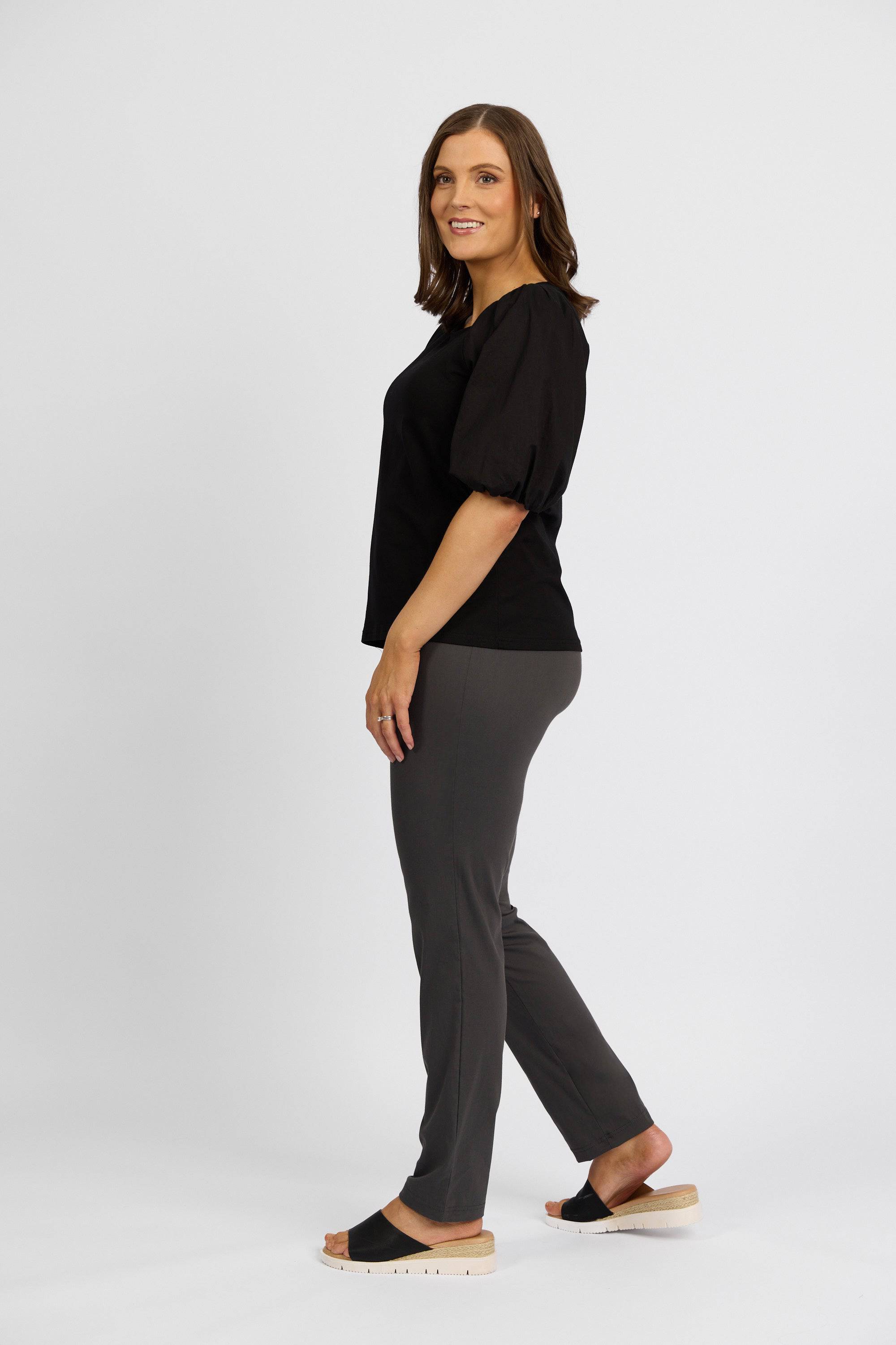 A woman with shoulder-length brown hair stands sideways, smiling, wearing a black short-sleeve blouse, Vassalli Paris Full Length Pant in Beluga gray stretch fabric, and black slide sandals with a light sole against a plain white background.