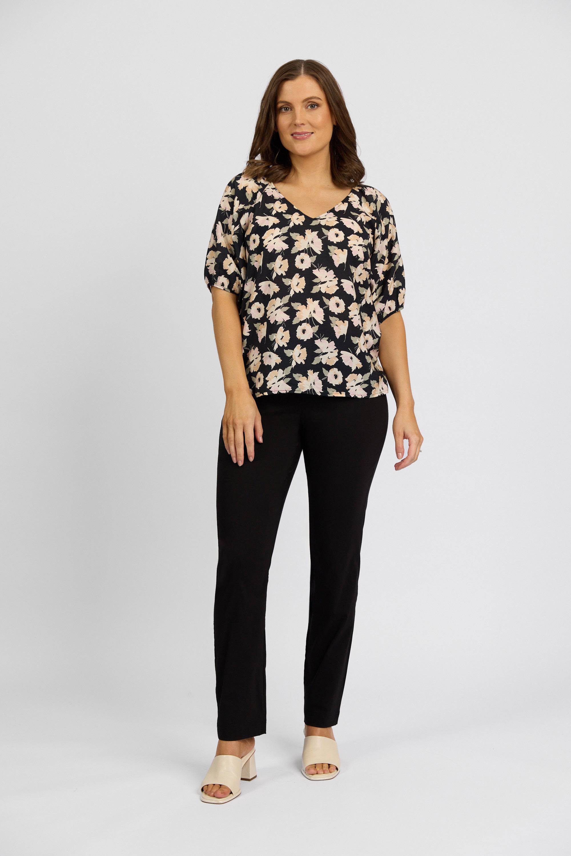 A woman stands facing forward wearing a black stretch top with a white floral pattern, Vassalli Paris Full Length Pant in black, and beige open-toe heels against a plain light background.