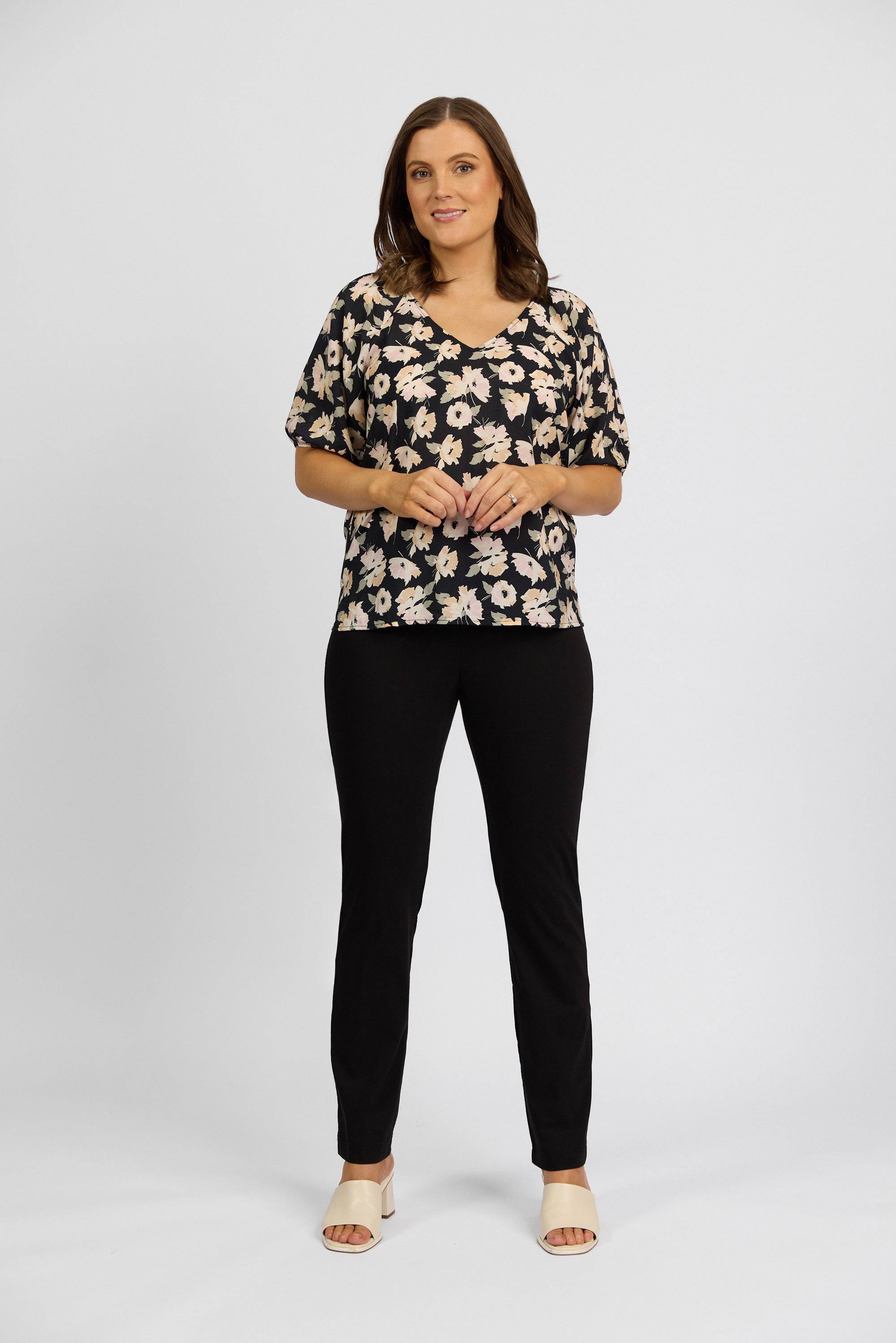 A woman stands against a white background wearing a black floral stretch short-sleeve top, Vassalli Paris Full Length Pant in black, and cream open-toe shoes. She has shoulder-length brown hair, smiles softly, and clasps her hands.