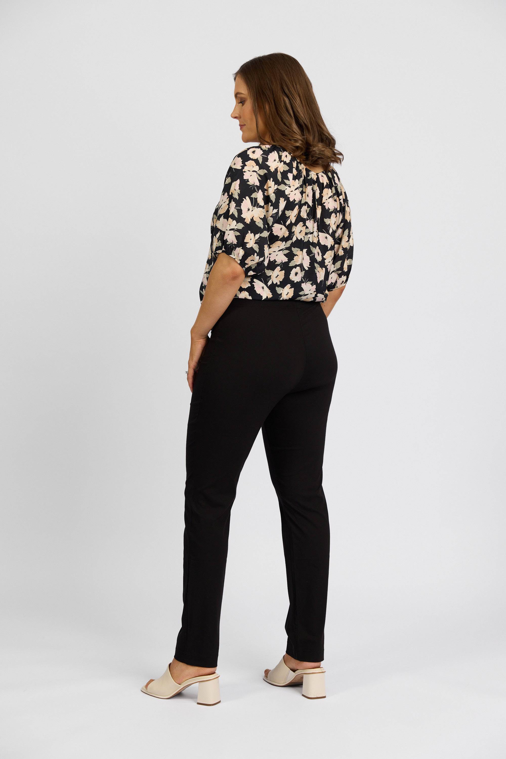A woman faces away, wearing a black floral blouse, Vassalli Paris Full Length Pant in black, and light-colored heeled shoes against a plain white background.
