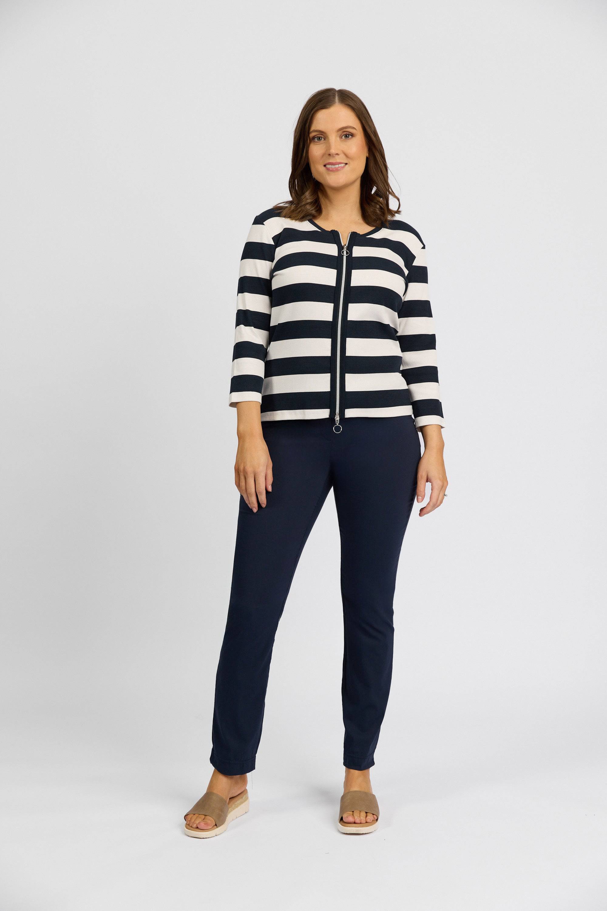 A woman stands facing forward, wearing a black and white striped zip-up sweater, Vassalli Paris Full Length Pant - Inka in dark navy stretch fabric, and light-colored sandals against a plain white background.