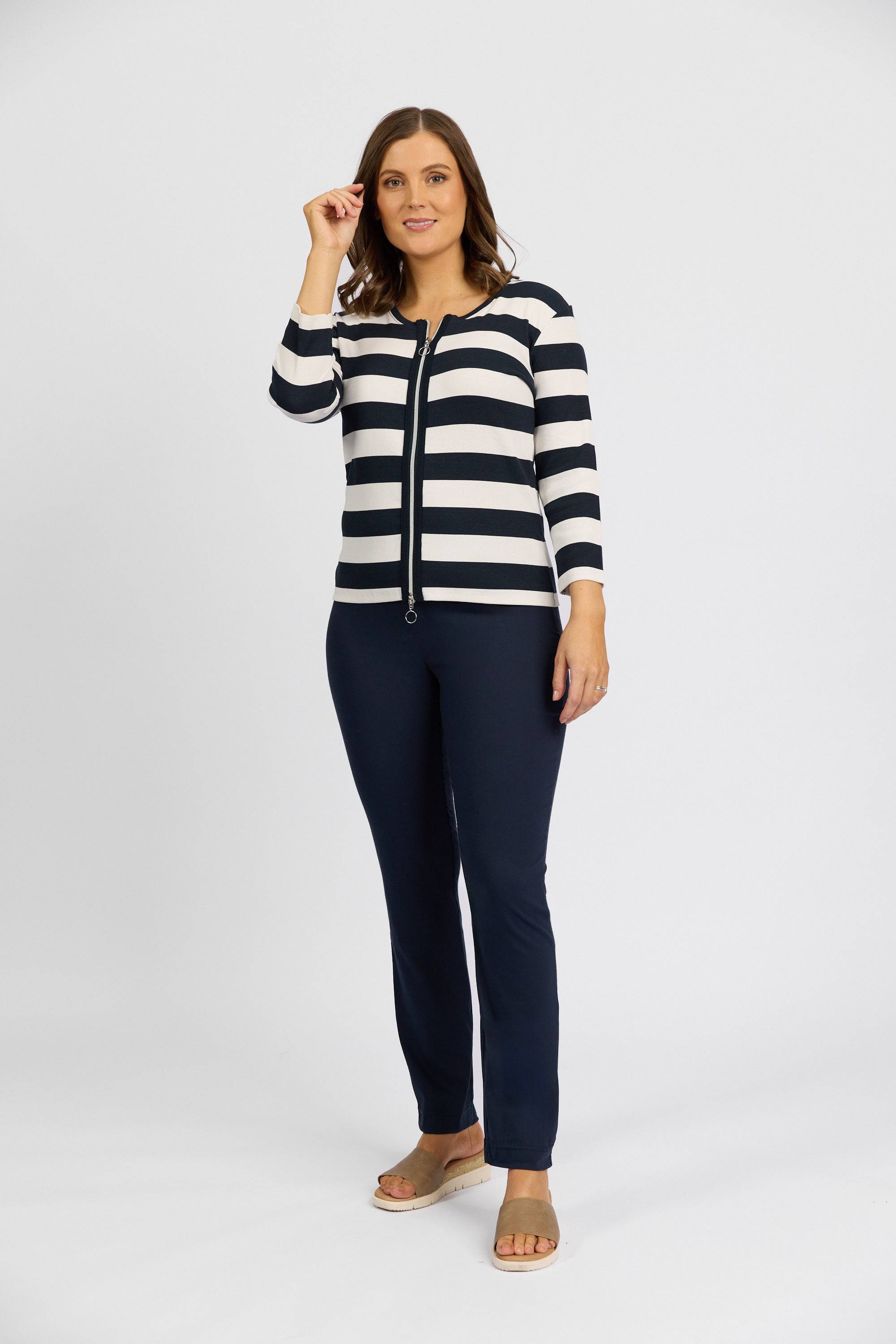 A woman stands against a plain light background, smiling slightly. She wears a navy and white striped long-sleeve top, Vassalli Paris Full Length Pant - Inka in dark blue stretch fabric, beige sandals, and her brown shoulder-length hair frames her face.
