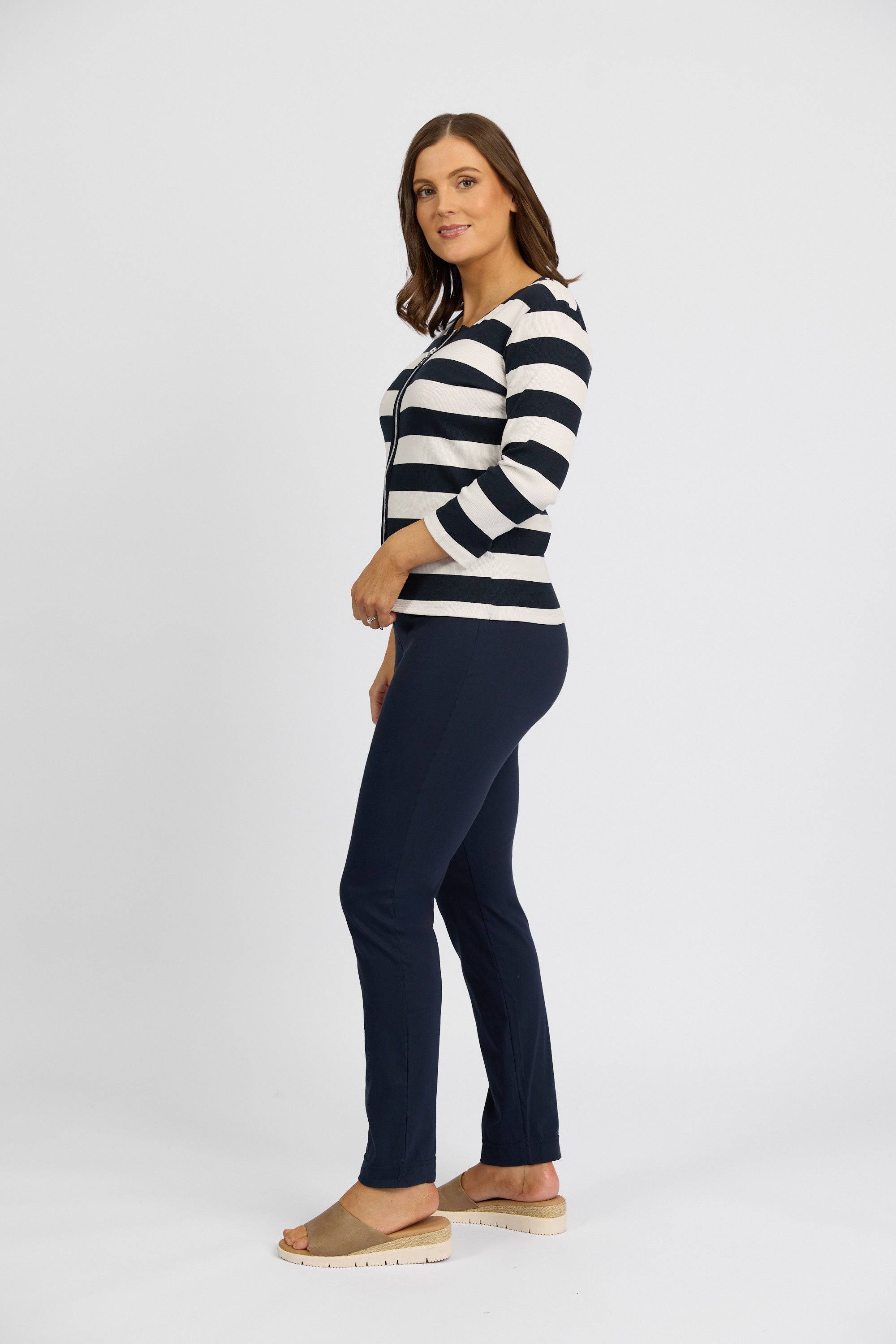 A woman with shoulder-length brown hair and a slight smile stands sideways against a white background, wearing Vassalli's Paris Full Length Pant - Inka in dark blue stretch fabric, a black and white striped long-sleeve top, and beige sandals.