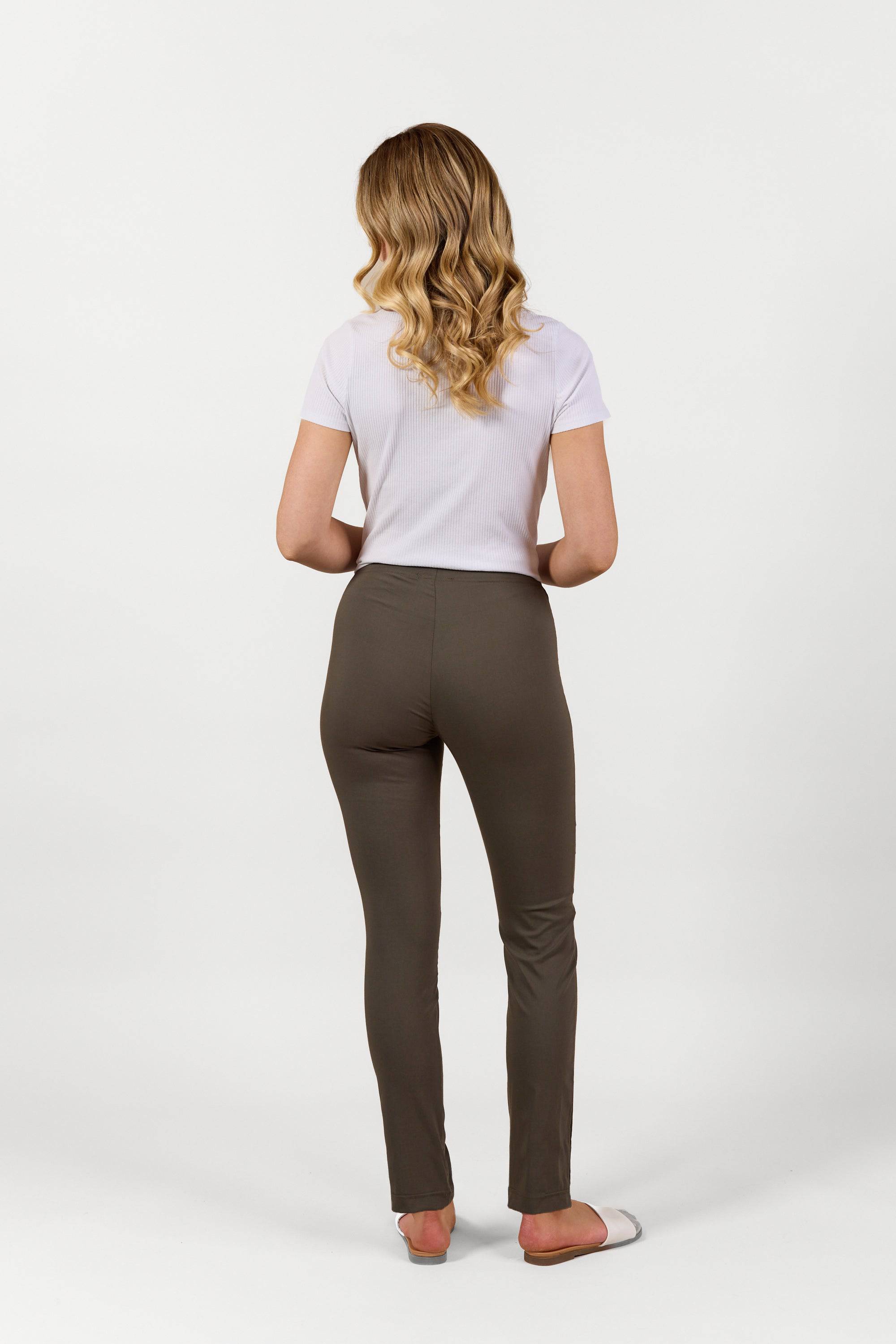 A woman with wavy blonde hair, facing away, wears a white short-sleeved shirt and Vassalli Paris Full Length Pant - Khaki, paired with white shoes against a plain white background.