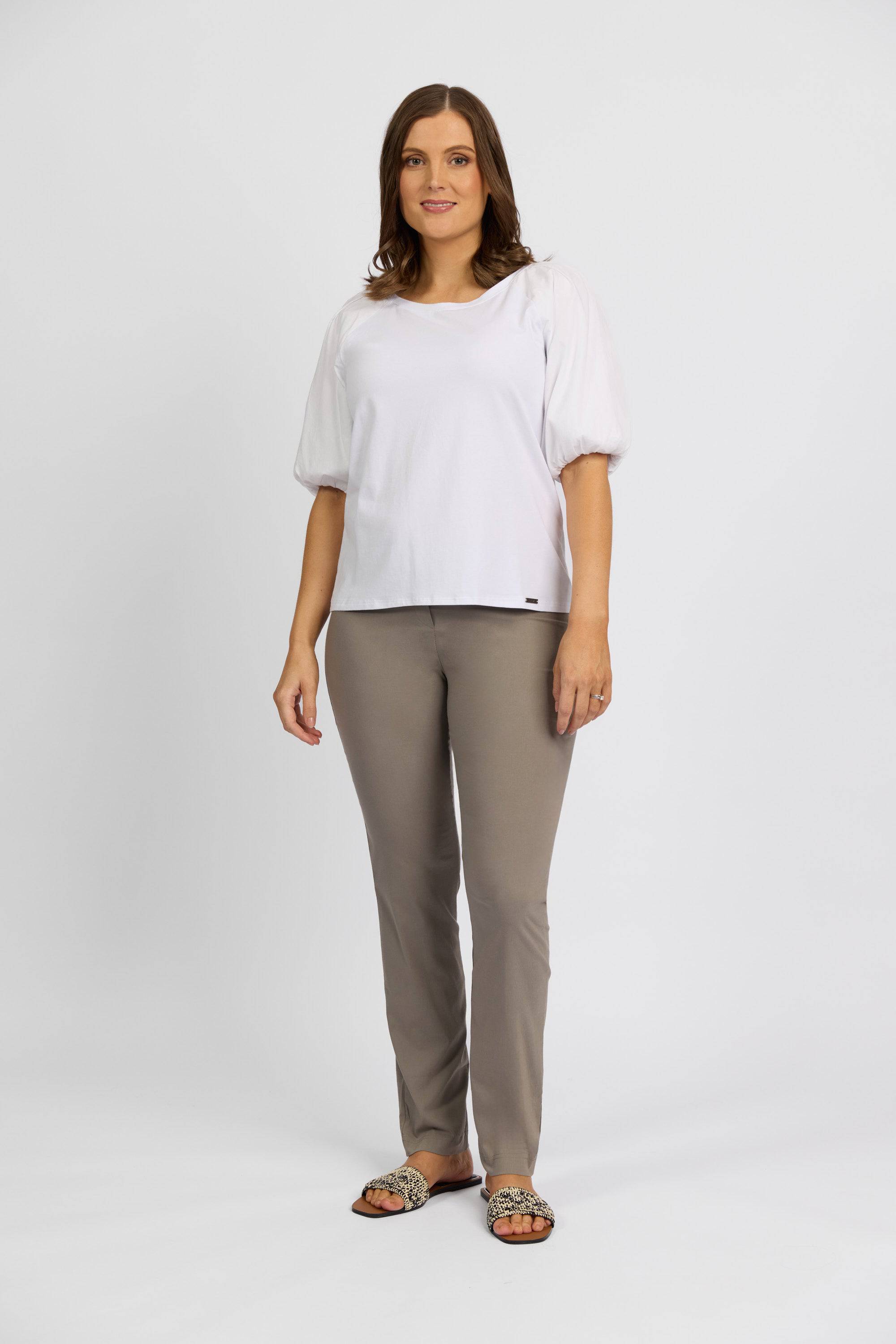 A woman with brown hair stands against a white background, wearing a white short-sleeved blouse, Vassalli Paris Full Length Pant in Latte, and animal print slide sandals. She smiles gently and has a relaxed posture.