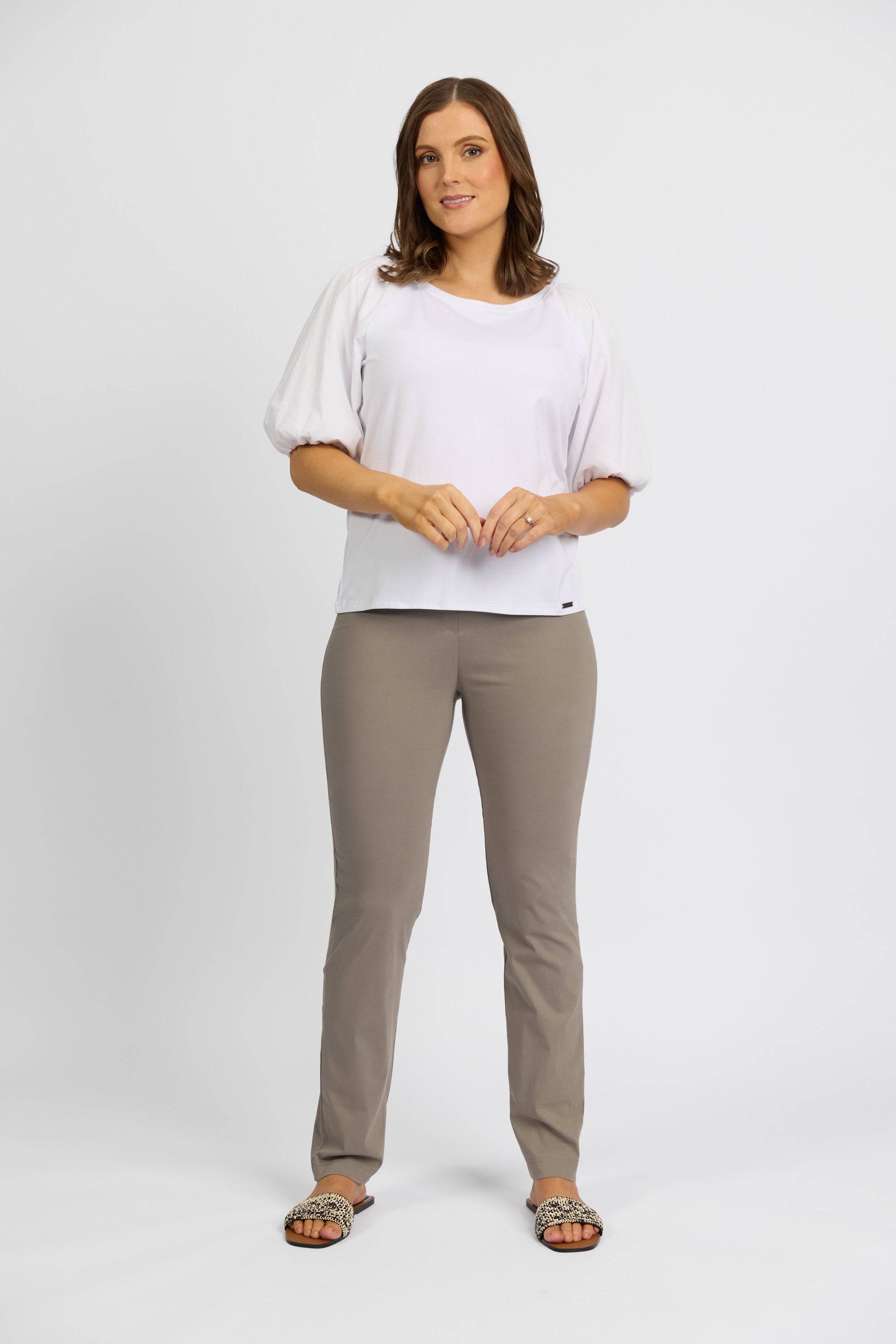 A woman with shoulder-length brown hair and a neutral expression wears a white blouse with puff sleeves, Vassalli's Paris Full Length Pant in Latte, and open-toed sandals against a plain white background.
