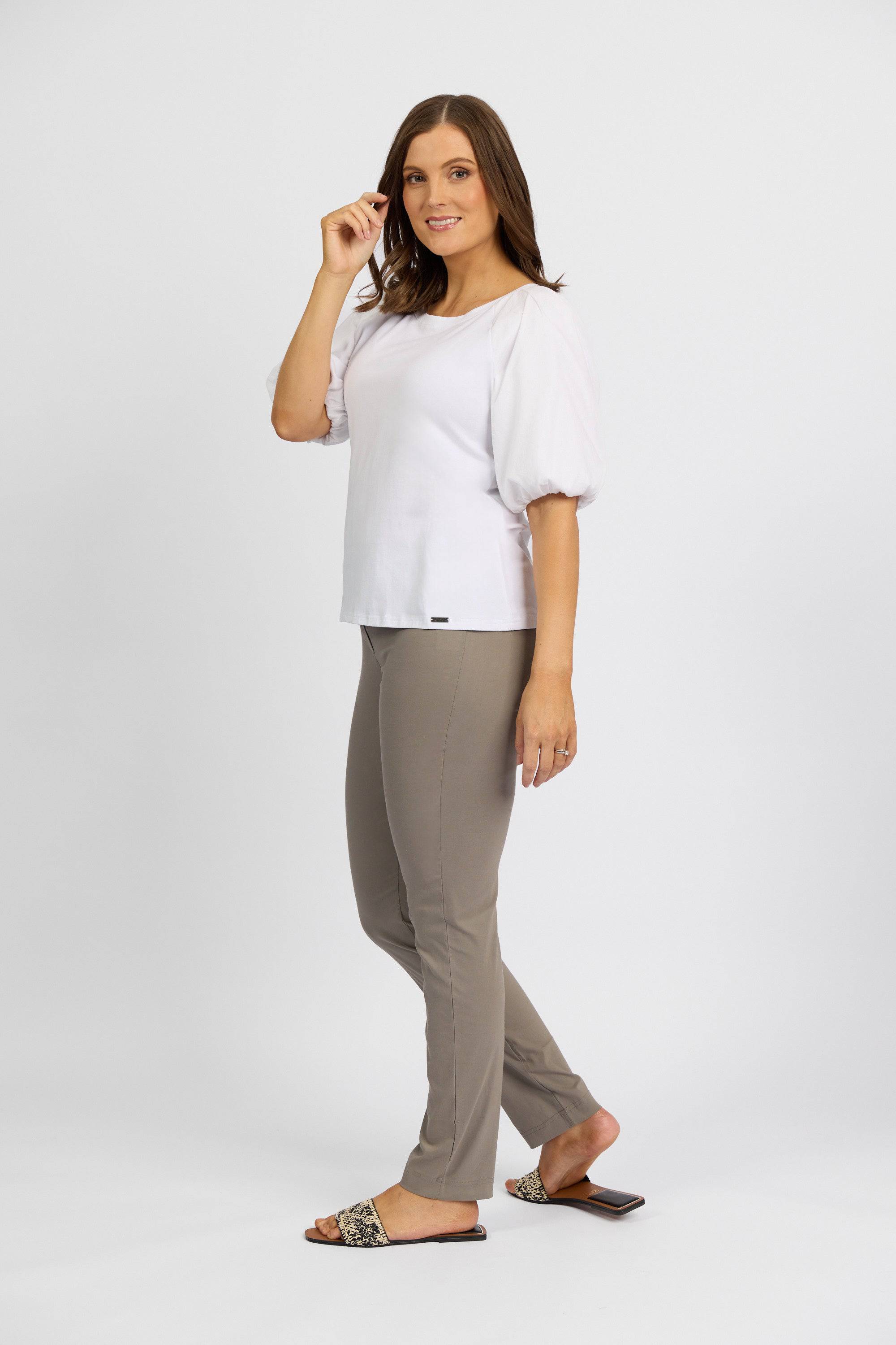 A woman with long brown hair smiles and touches her hair against a plain white background. She wears a white puff-sleeve top and Vassalli’s Paris Full Length Pant in Latte, paired with patterned slide sandals.