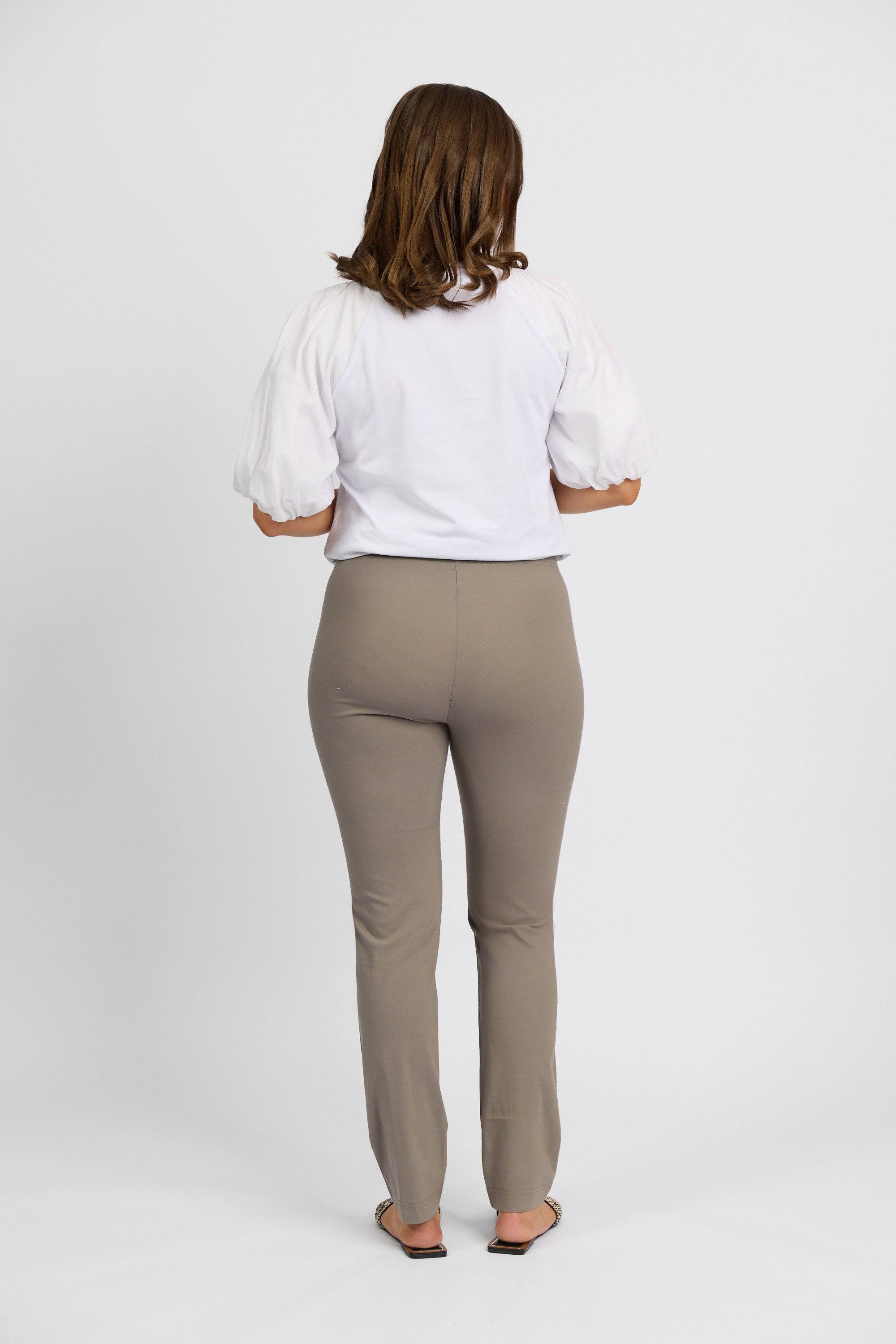 A person with shoulder-length brown hair, wearing a white short-sleeve top and Vassalli Paris Full Length Pant - Latte, stands barefoot facing away from the camera against a plain white background.
