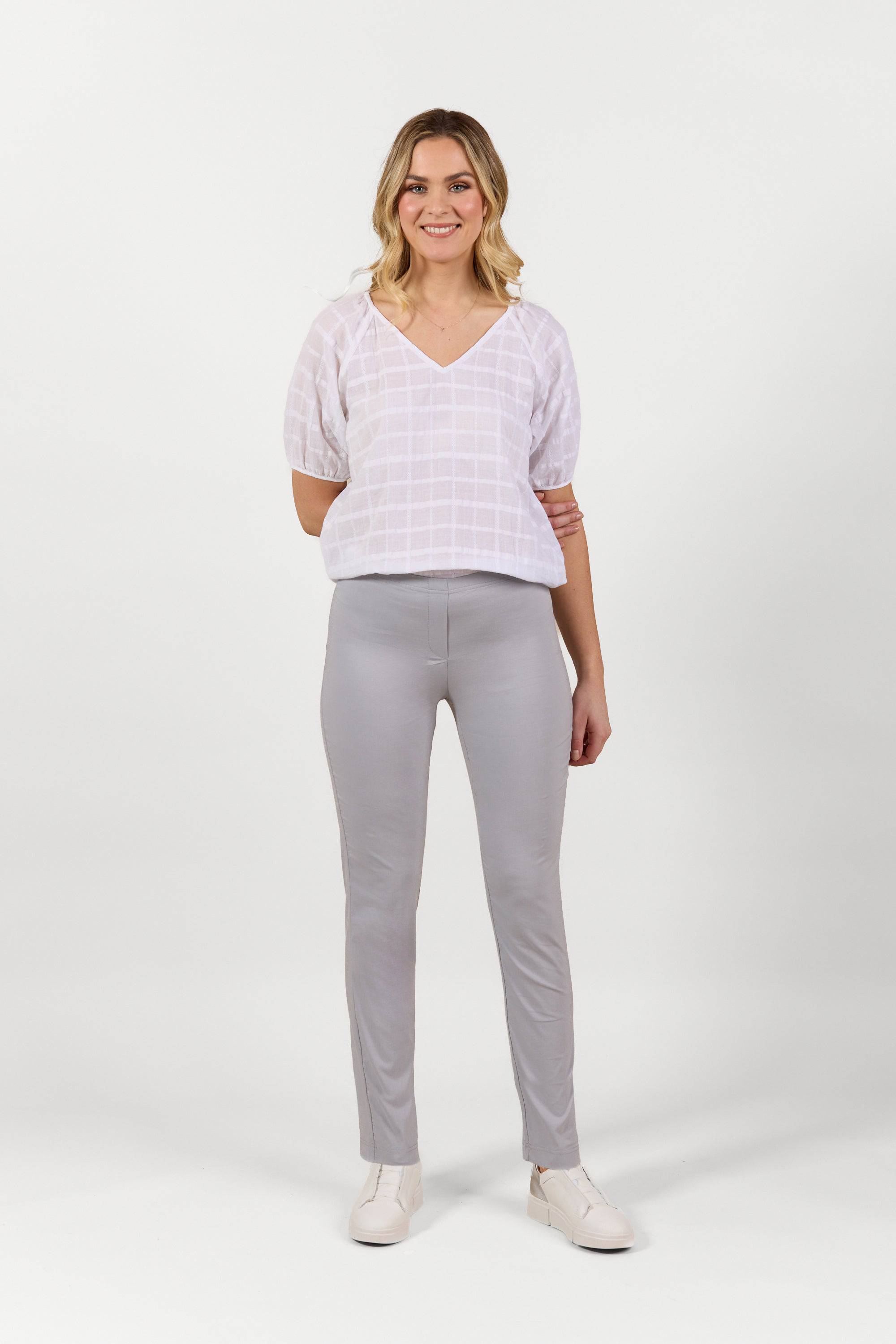 A woman with shoulder-length blonde hair smiles against a white background, wearing a light pink checkered blouse, Vassalli Paris Full Length Pant in Silver, and white shoes. Her arms are relaxed by her sides.