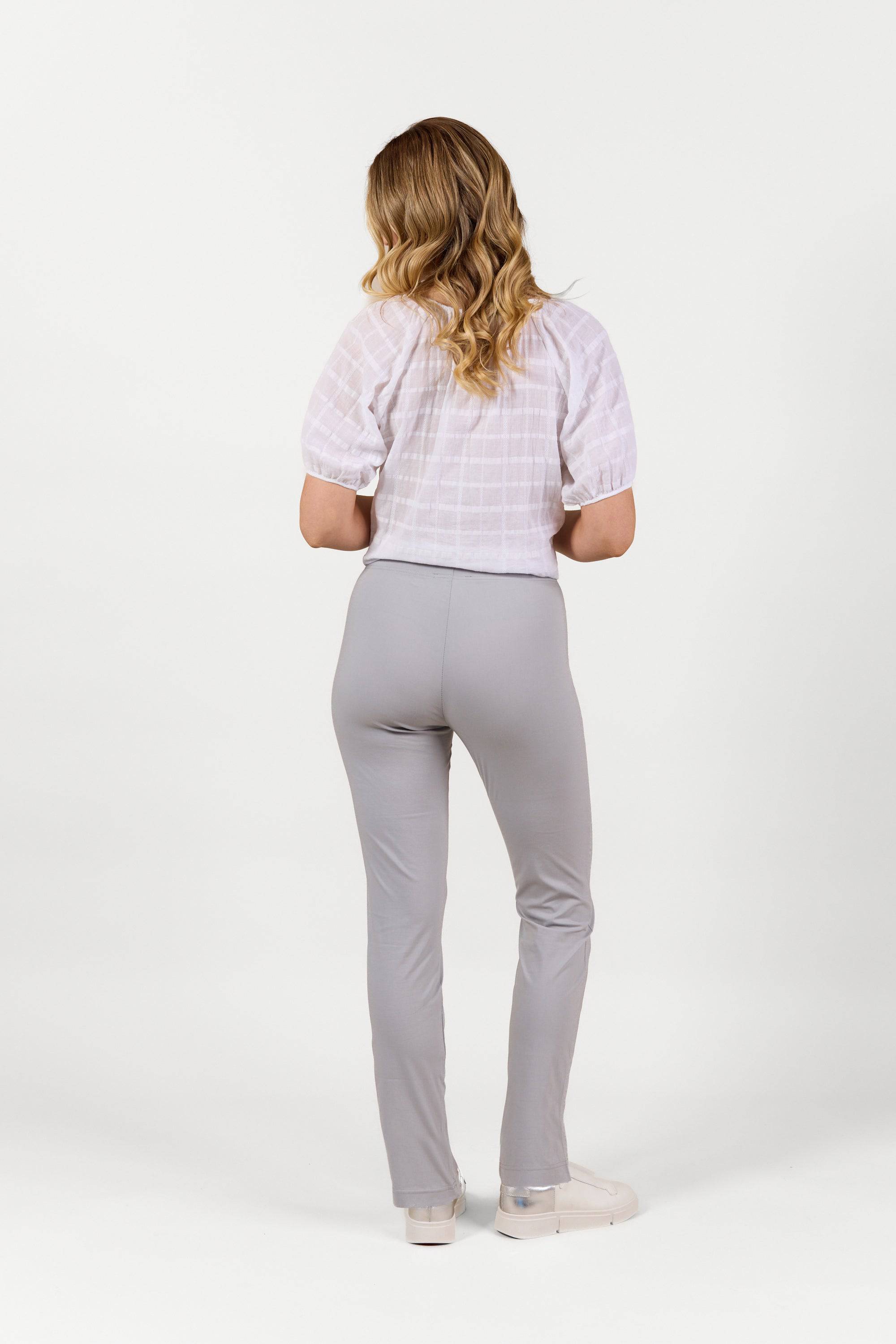 A woman with wavy blonde hair, wearing a white short-sleeve blouse and Vassalli Paris Full Length Pant in Silver, stands facing away against a plain white background.