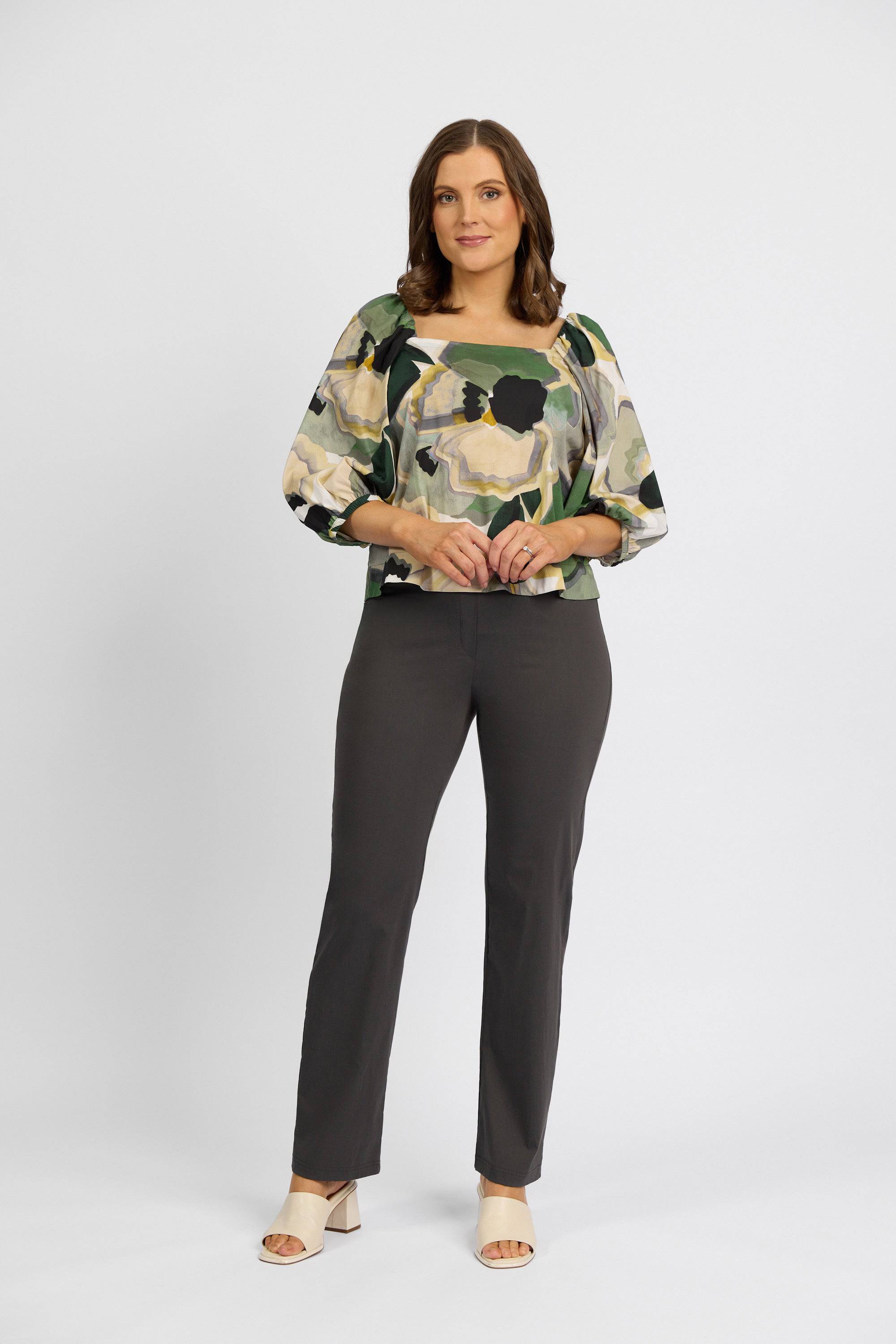 A woman with shoulder-length brown hair and a relaxed expression stands against a white background, wearing a floral blouse, beige open-toe heels, and Vassalli’s Paris Full Length Pant - Beluga in dark gray.