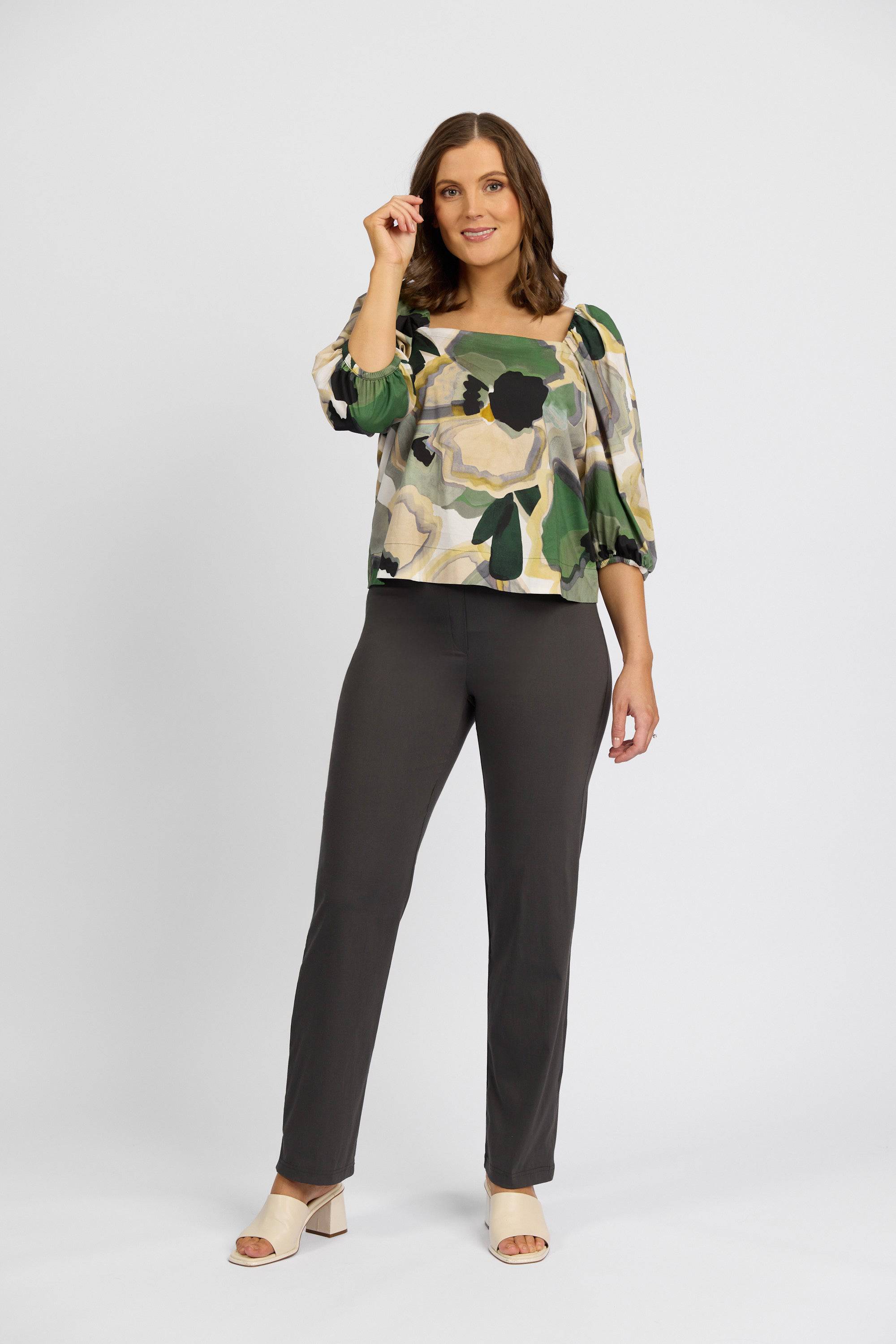 A woman stands against a plain white background, wearing a patterned blouse with green, beige, and black hues. She pairs it with Vassalli's Paris Full Length Pant - Beluga and light open-toe heels, smiling as she touches her hair.