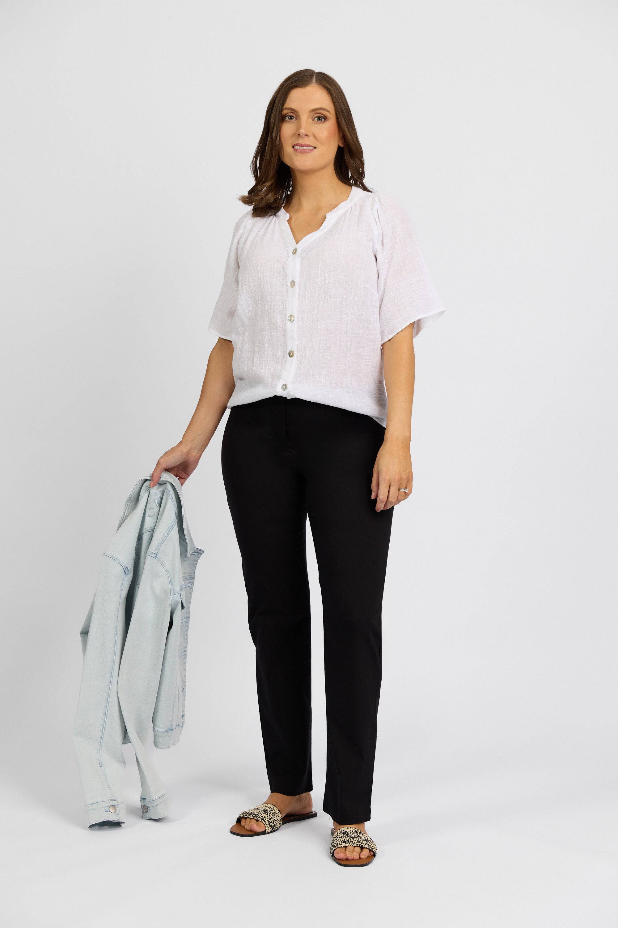 A woman stands against a plain white background, wearing a white short-sleeve button-up blouse, Vassalli Paris Full Length Pant in black, patterned slide sandals, and holds a light blue denim jacket in her left hand while smiling slightly.