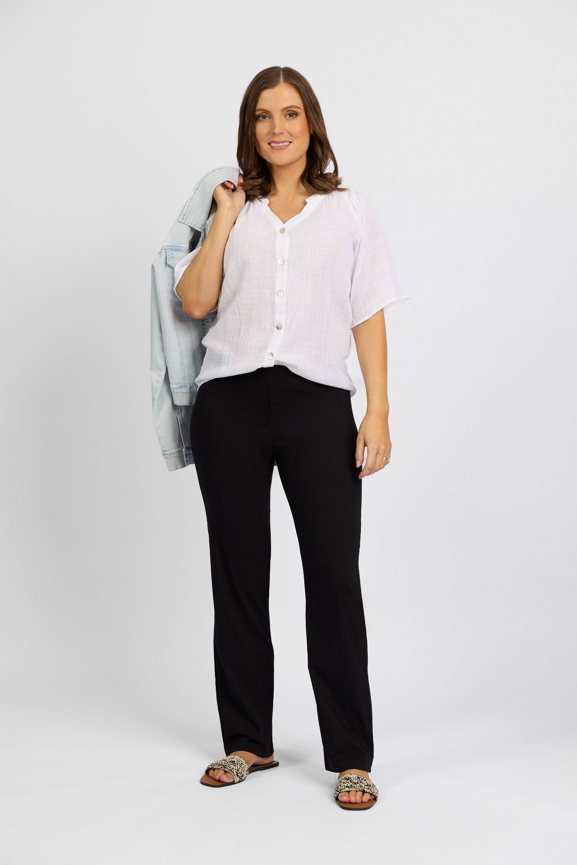 A woman smiles at the camera wearing a white short-sleeve blouse, Vassalli Paris Full Length Pant in black, and leopard-print sandals. She stands against a plain background, holding a light blue denim jacket over her shoulder.