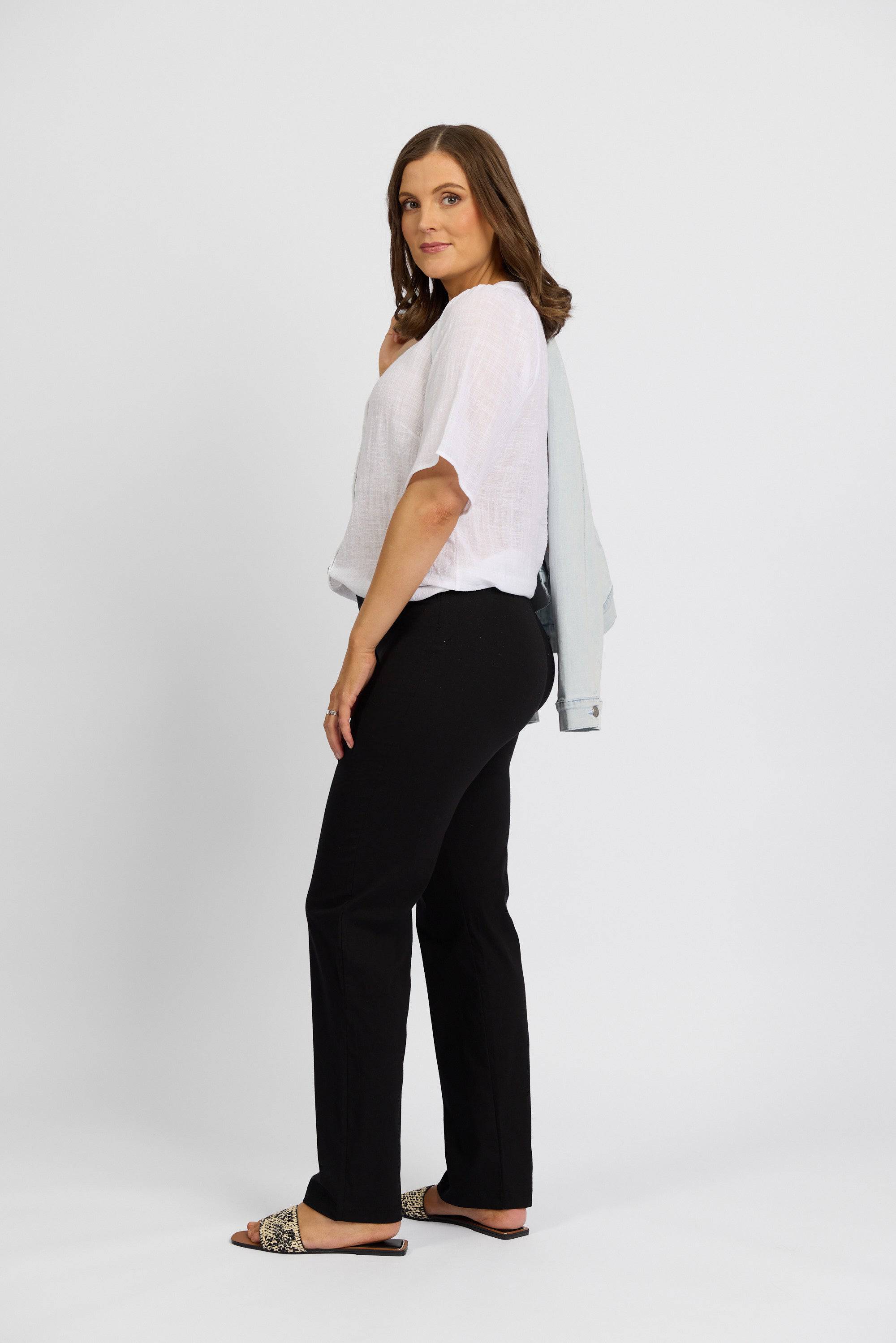 A woman stands sideways against a plain white background wearing a white short-sleeve blouse, Vassalli Paris Full Length Pant - Black, and leopard print slide sandals, with a light jacket draped over her shoulder, looking at the camera.