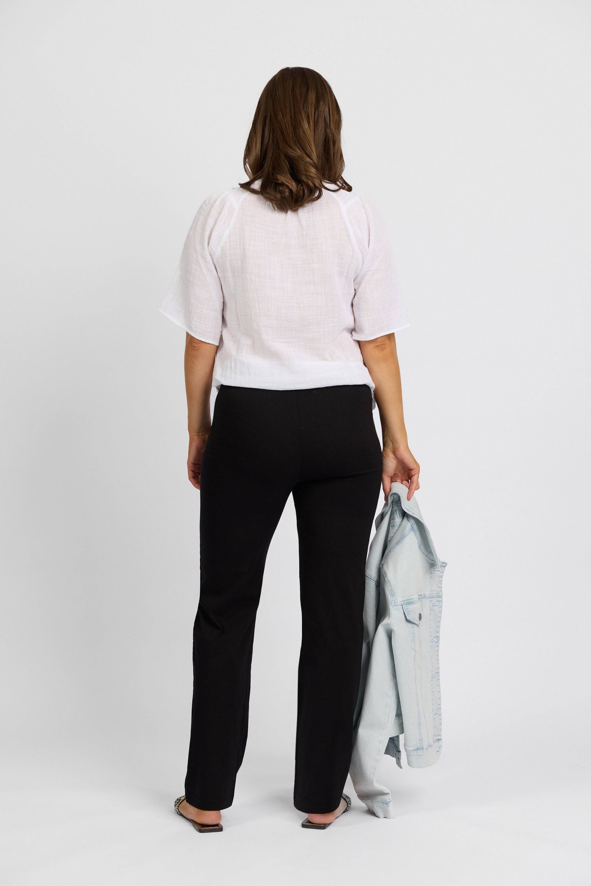 A woman stands facing away in a white short-sleeve blouse and Vassalli Paris Full Length Pant - Black, crafted from stretch fabric. She holds a light blue denim jacket in her left hand against a plain white background.