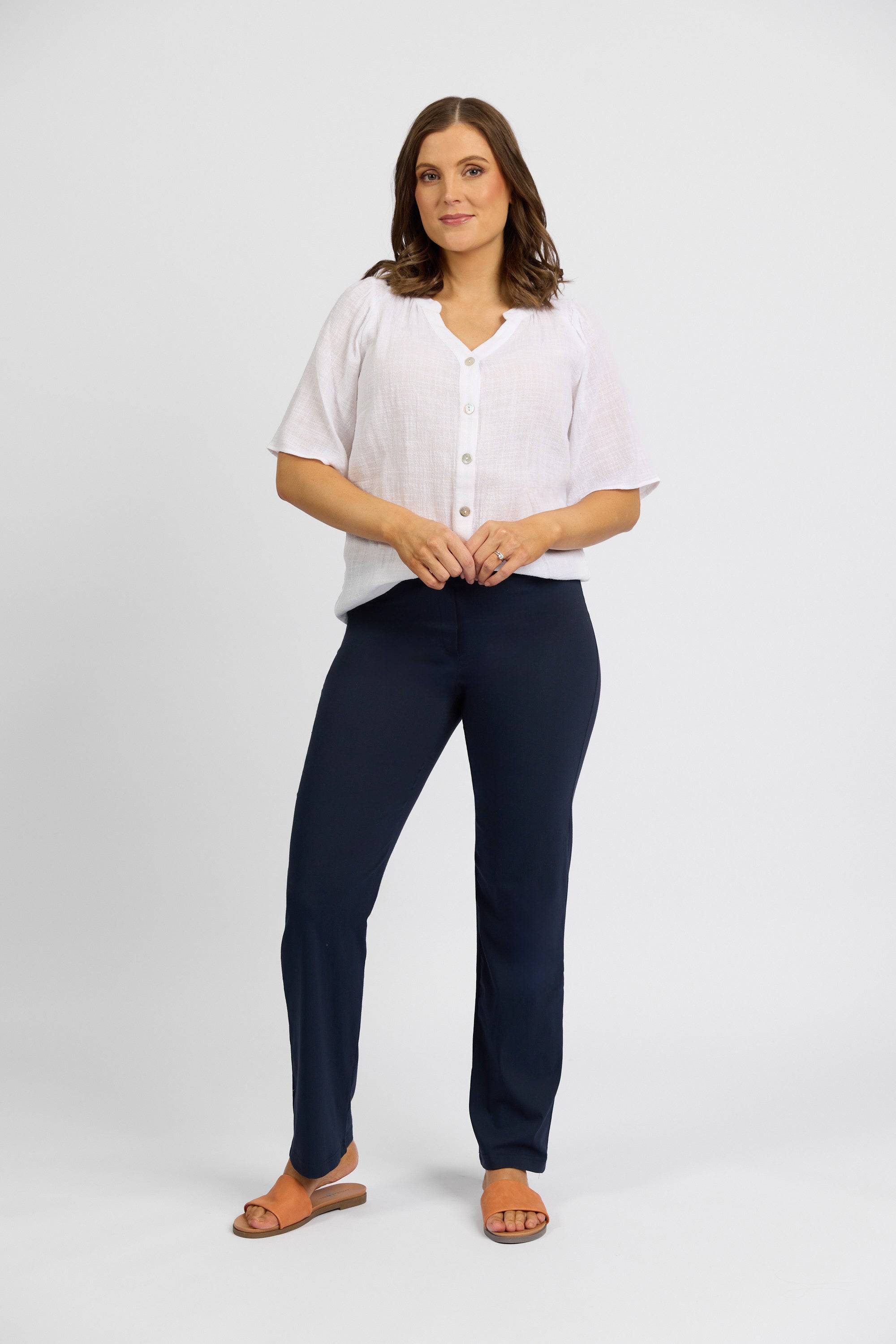 A woman with shoulder-length brown hair and a neutral expression poses against a white background, wearing Vassalli Paris Full Length Pant - Inka in navy blue, a white short-sleeve button-up blouse, and orange sandals.